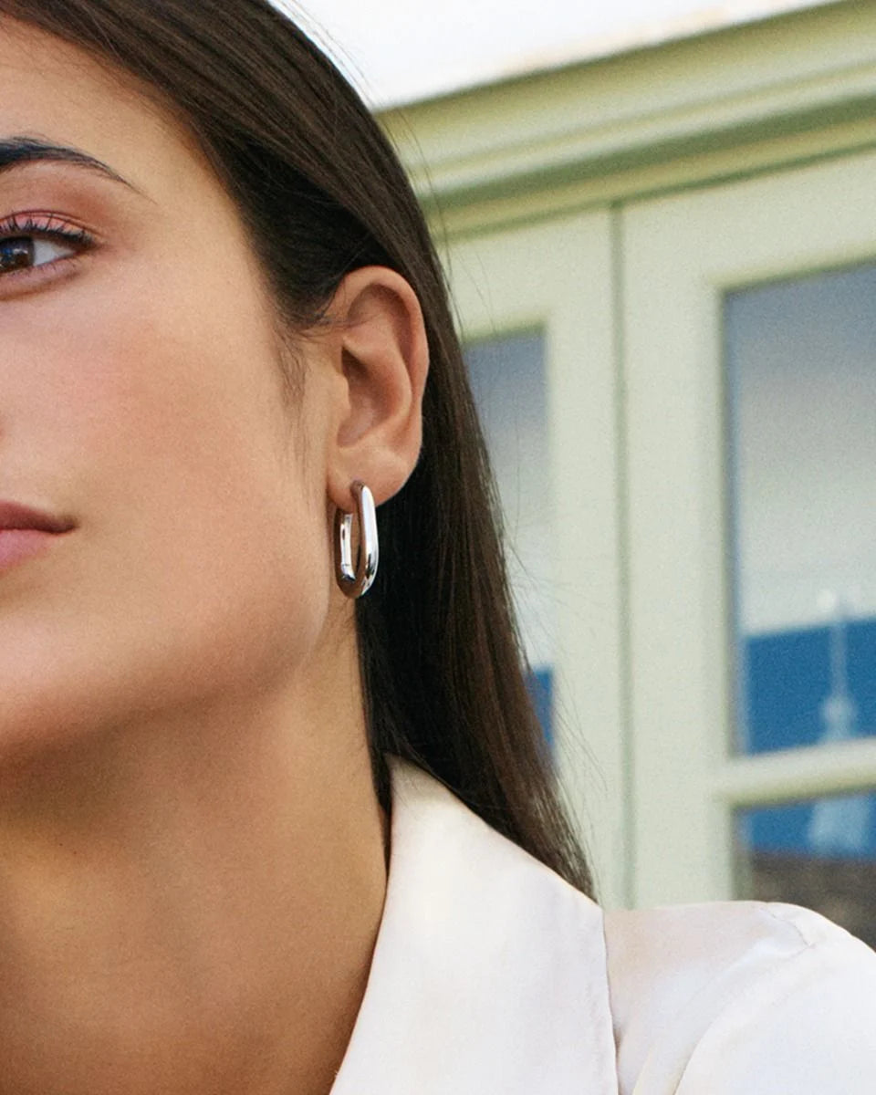 Close-up of a person with long brown hair, wearing EDBLAD Modernist Hoops L and a white shirt, standing in front of a light green window frame.