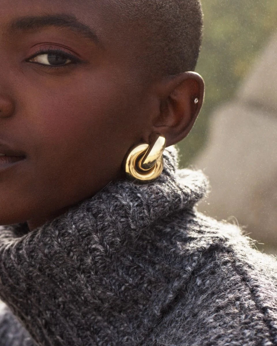 Close-up of a person wearing a gray textured sweater and EDBLAD's Redondo Studs L in 14K gold plating. The background is blurred.