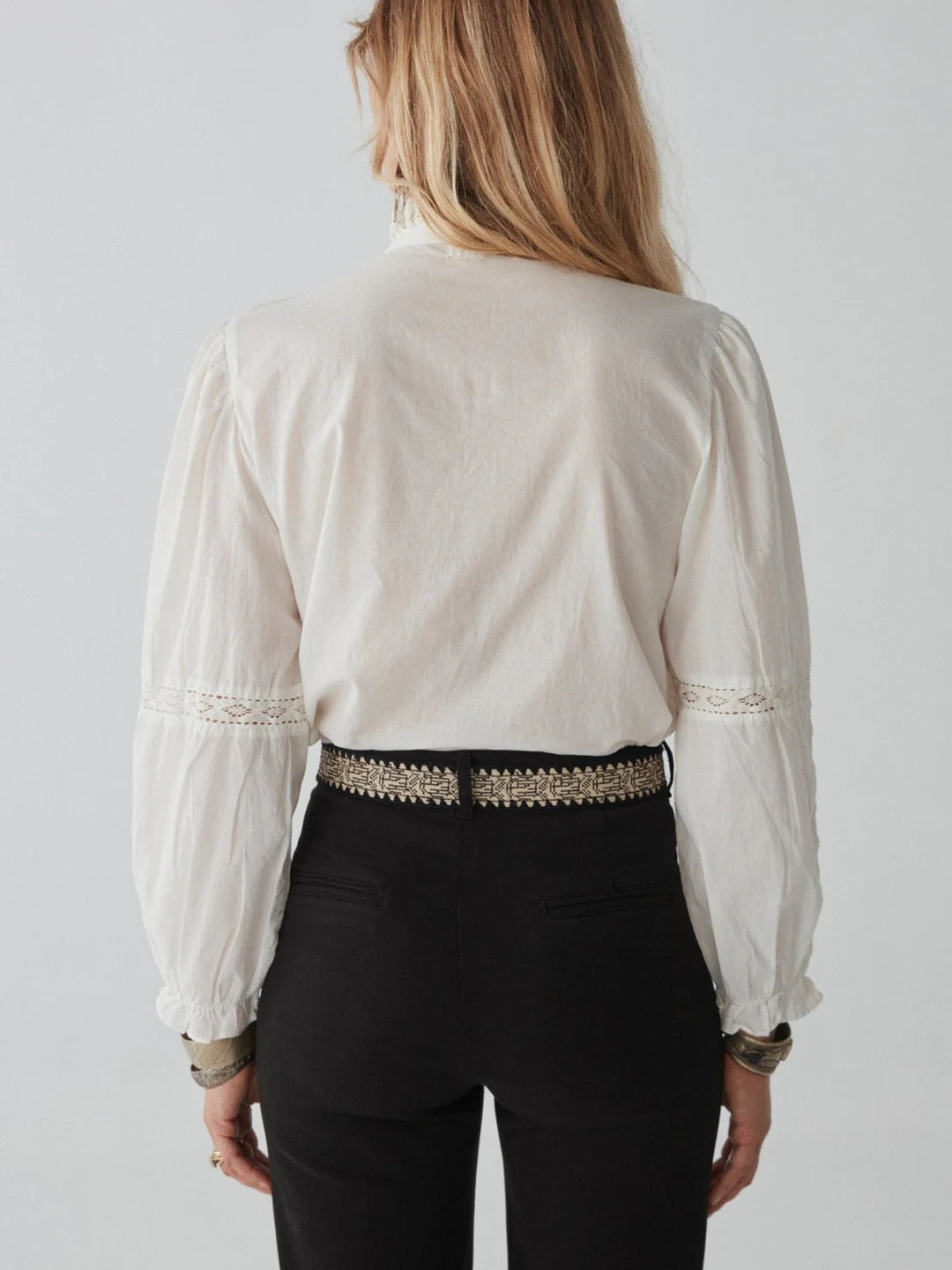 A person with long hair is seen from the back, wearing the Emanuelle Blouse in white by Maison Hotel, adorned with lace accents, paired with high-waisted black pants featuring a decorative belt.