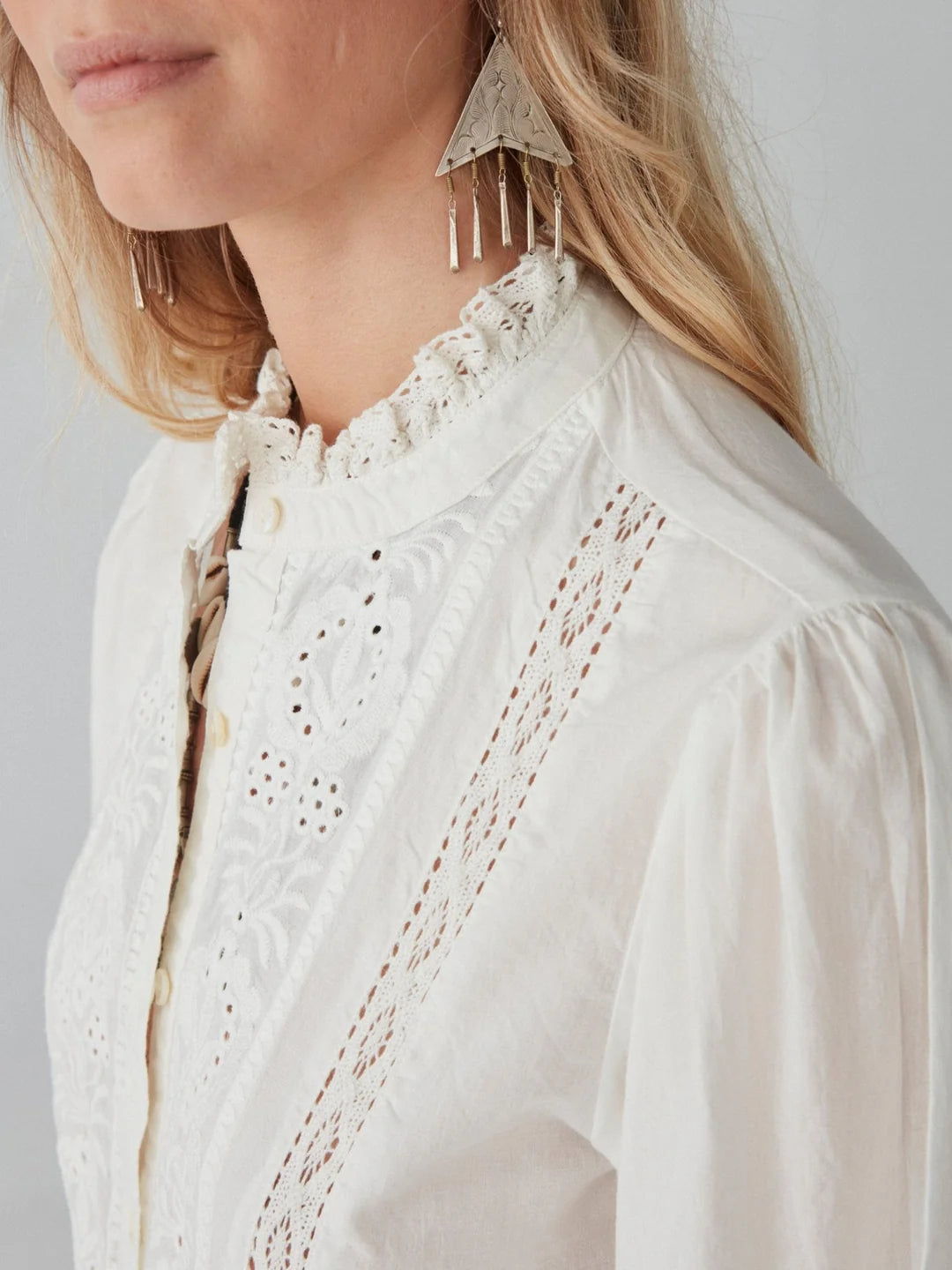 A person wearing the Emanuelle Blouse - White by Maison Hotel, featuring a high collar and lace accents, complemented with triangular earrings.