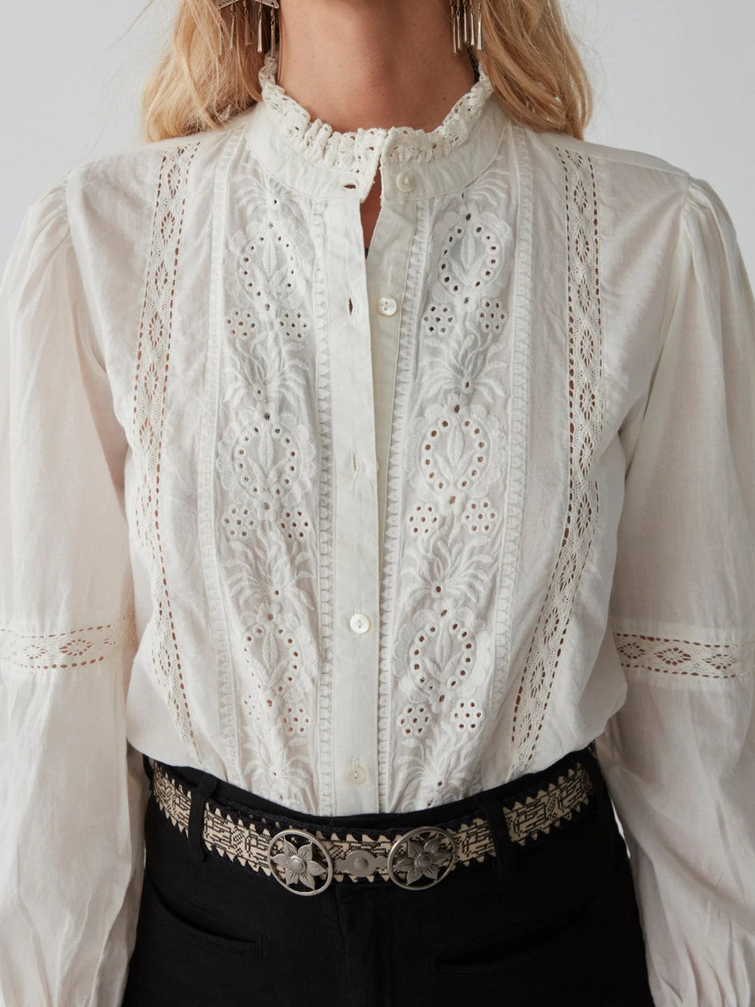 A person wearing the Emanuelle Blouse in white by Maison Hotel, which features lace accents and a high collar. The blouse is elegantly paired with black pants adorned with an ornate silver belt. The image captures from shoulders to waist.