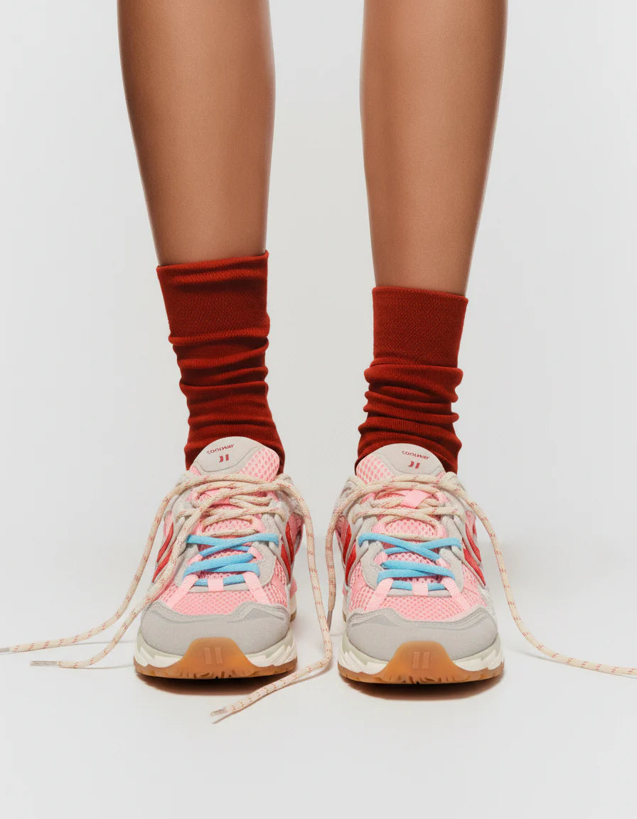 A person in red socks wears Coolway 2003 Sneakers - Pink Volt, featuring pink, blue, and gray suede leather with long, loosely tied laces, standing on a white background.