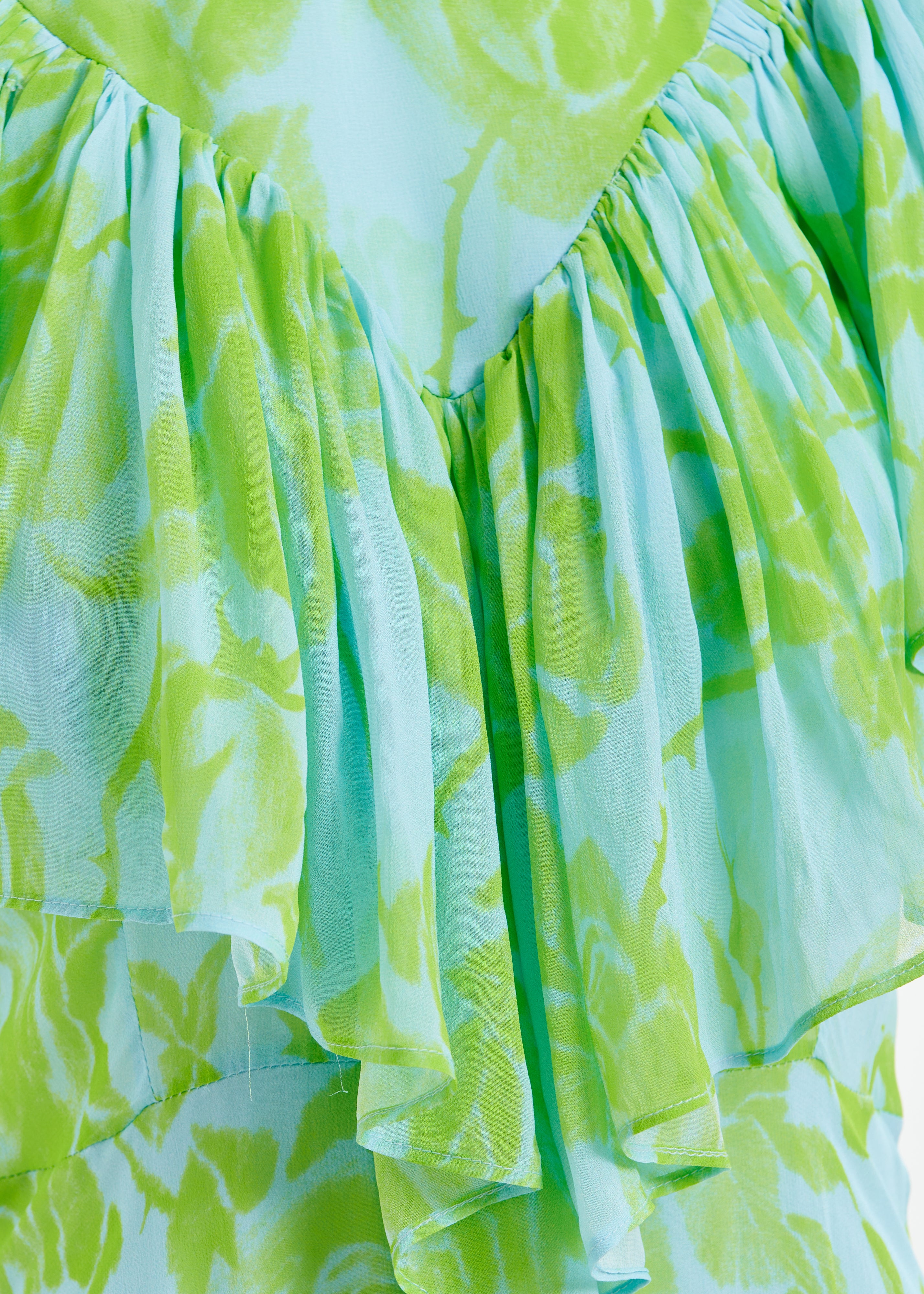 A close-up of the Honolulu Dress by Essentiel Antwerp reveals its green and light blue floral pattern, reminiscent of a relaxed fit dress with gathered pleats for a layered effect.