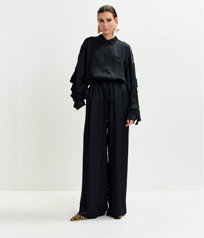 A person stands against a white background, wearing a loose black button-up shirt with ruffled sleeves, Essentiel Antwerp's Irving Wide Trousers in black featuring a high-rise waist, and leopard print shoes.