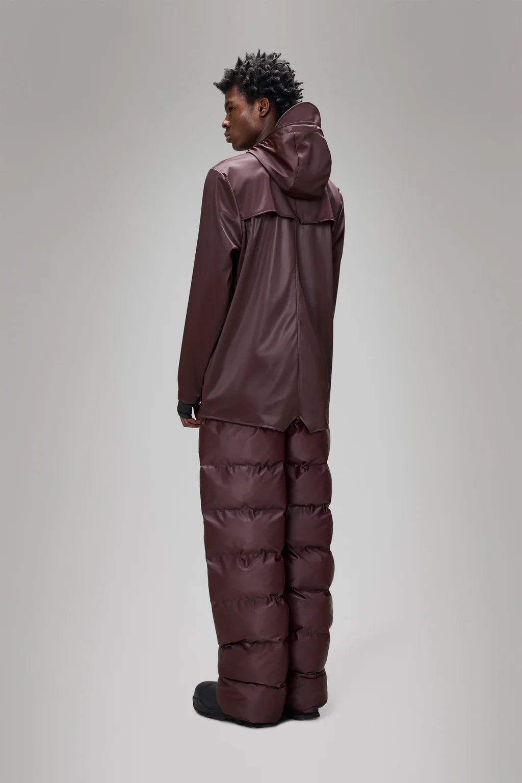 A person stands facing away, wearing the Rains Jacket W3 - Depth and matching puffer pants against a plain gray background.