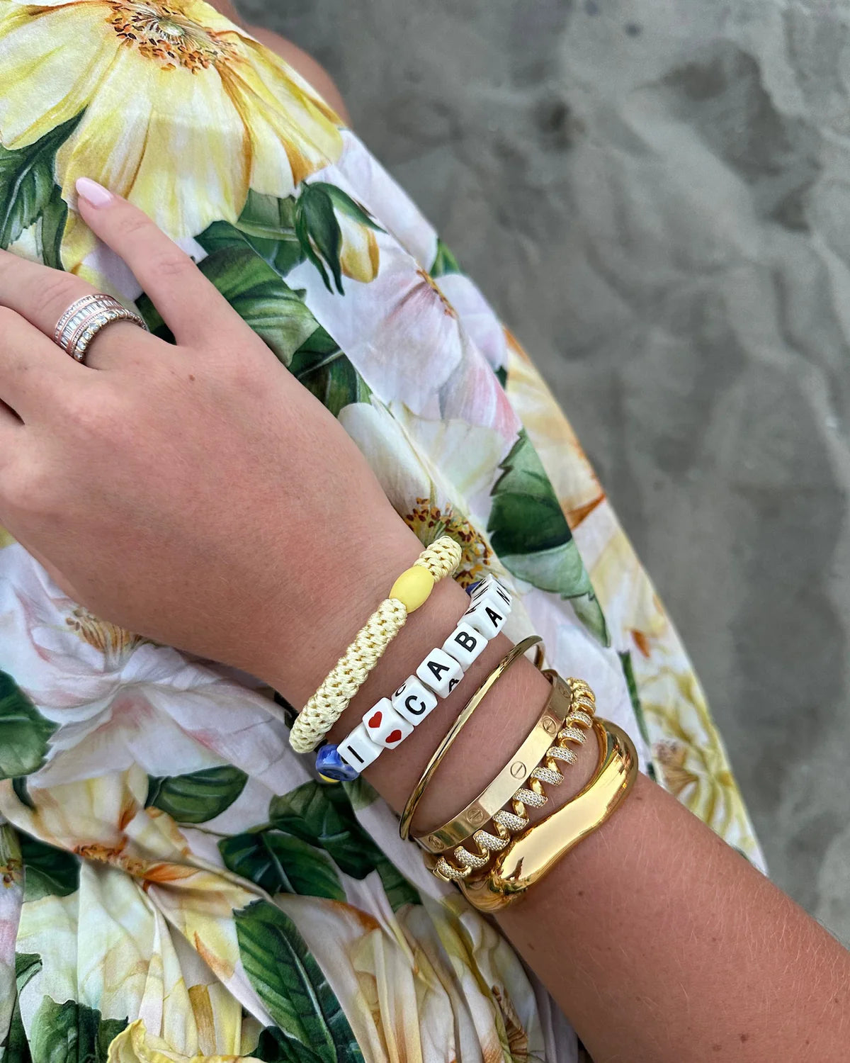 A hand wearing gold and beaded bracelets—including one with letter beads spelling "CABA"—and a KKNEKKI Hair Tie - Original rests on a yellow and green floral dress, with sand in the background.