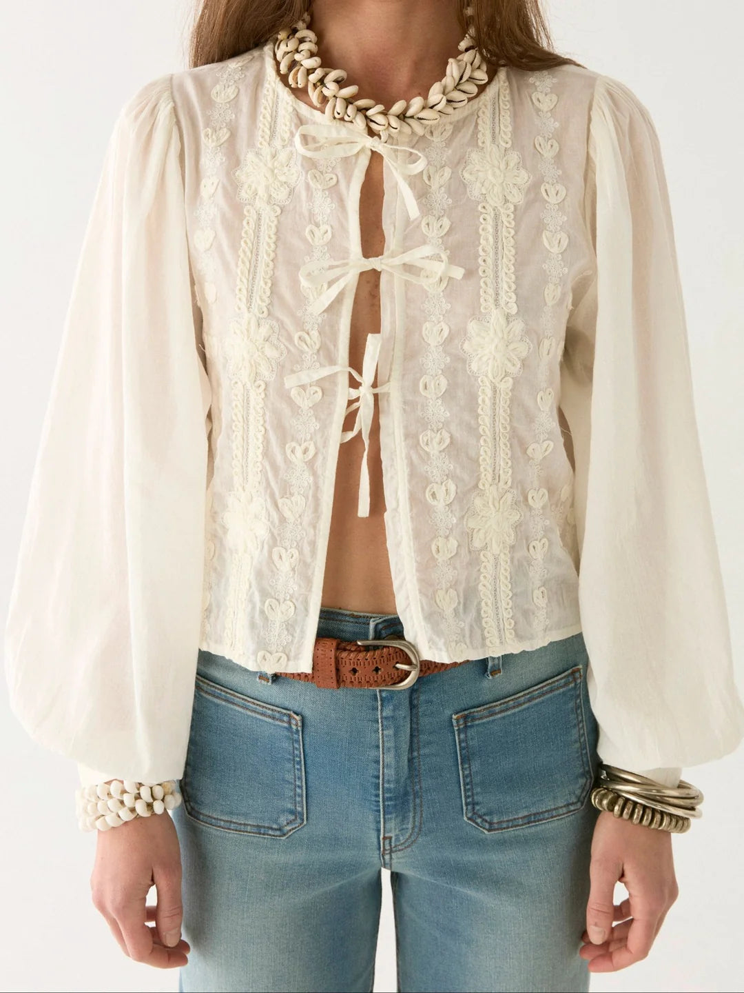 Someone in high-waisted jeans and an ivory Ludovica Blouse by Maison Hotel, featuring lace inserts and tie closures, accessorized with shell jewelry on the neck and wrists.