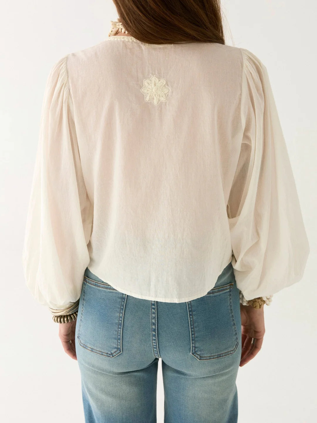 A person with long hair, facing away, is wearing Maison Hotel's Ludovica Blouse - Ivory, showcasing oversized puff sleeves and lace inserts. The blouse has floral details on the back and is paired effortlessly with blue jeans.