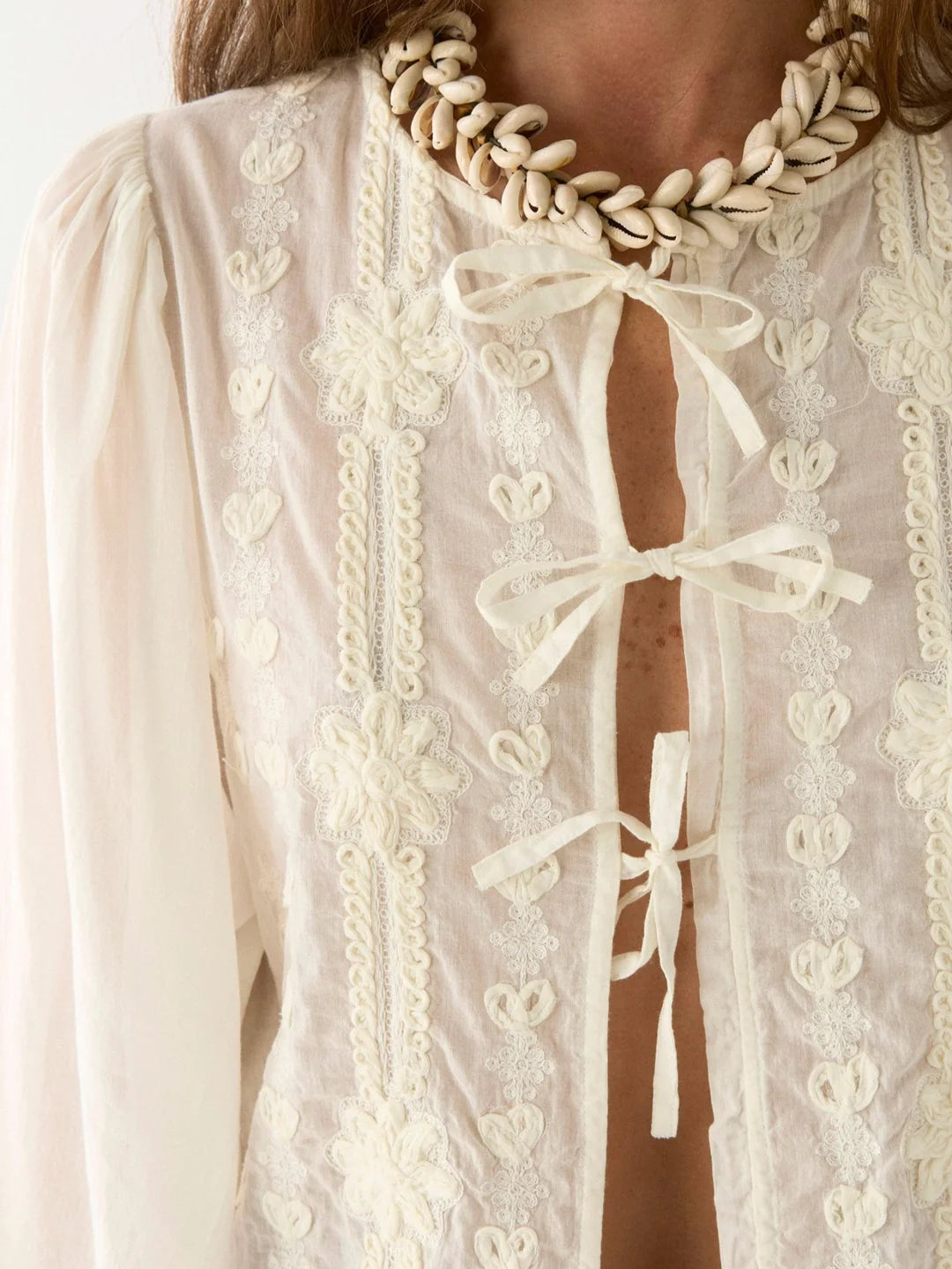 Someone is wearing the Maison Hotel's Ludovica Blouse - Ivory, which showcases oversized sleeves, an embroidered off-white design with bows, and pairs it with a shell necklace.