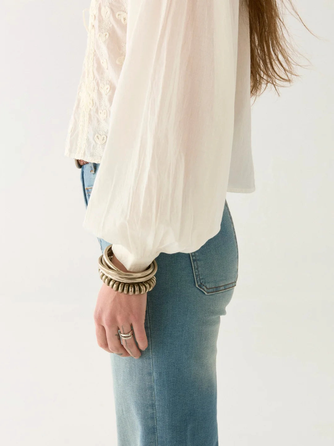 Wearing a Maison Hotel Ludovica Blouse in ivory with lace inserts and blue jeans, a person stands against a plain background, adorned with bracelets and rings on their left hand.