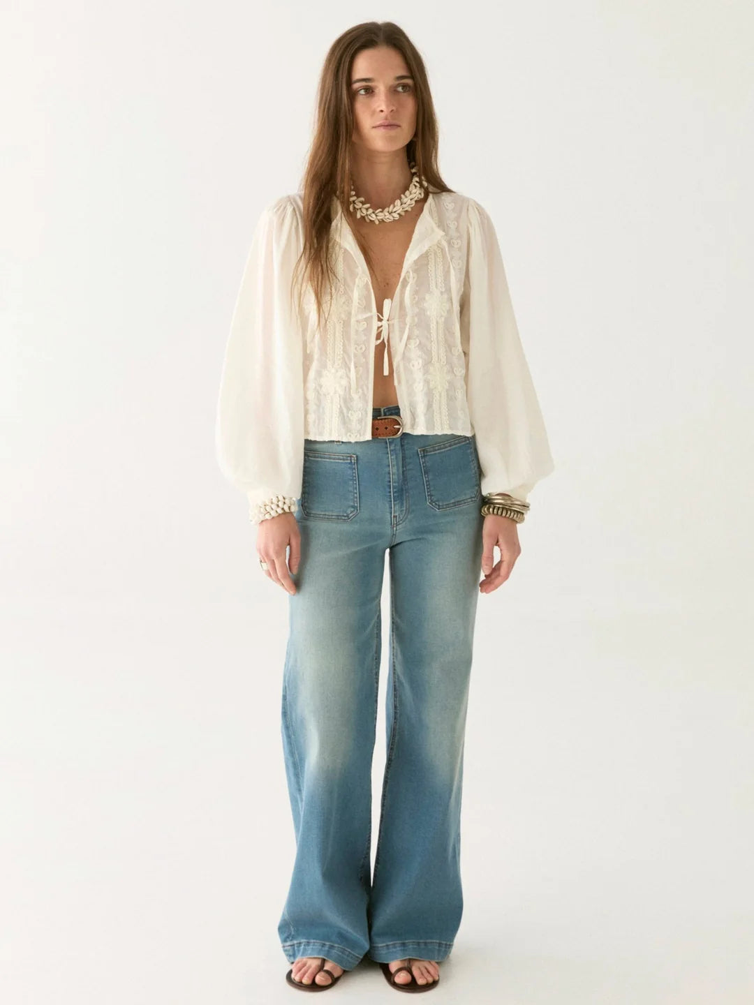 A person with long hair stands against a plain background wearing an elegant Ludovica Blouse - Ivory by Maison Hotel, its oversized sleeves adding drama. Paired with wide-legged jeans, a belt, and sandals, this look perfectly balances modern style with timeless grace.