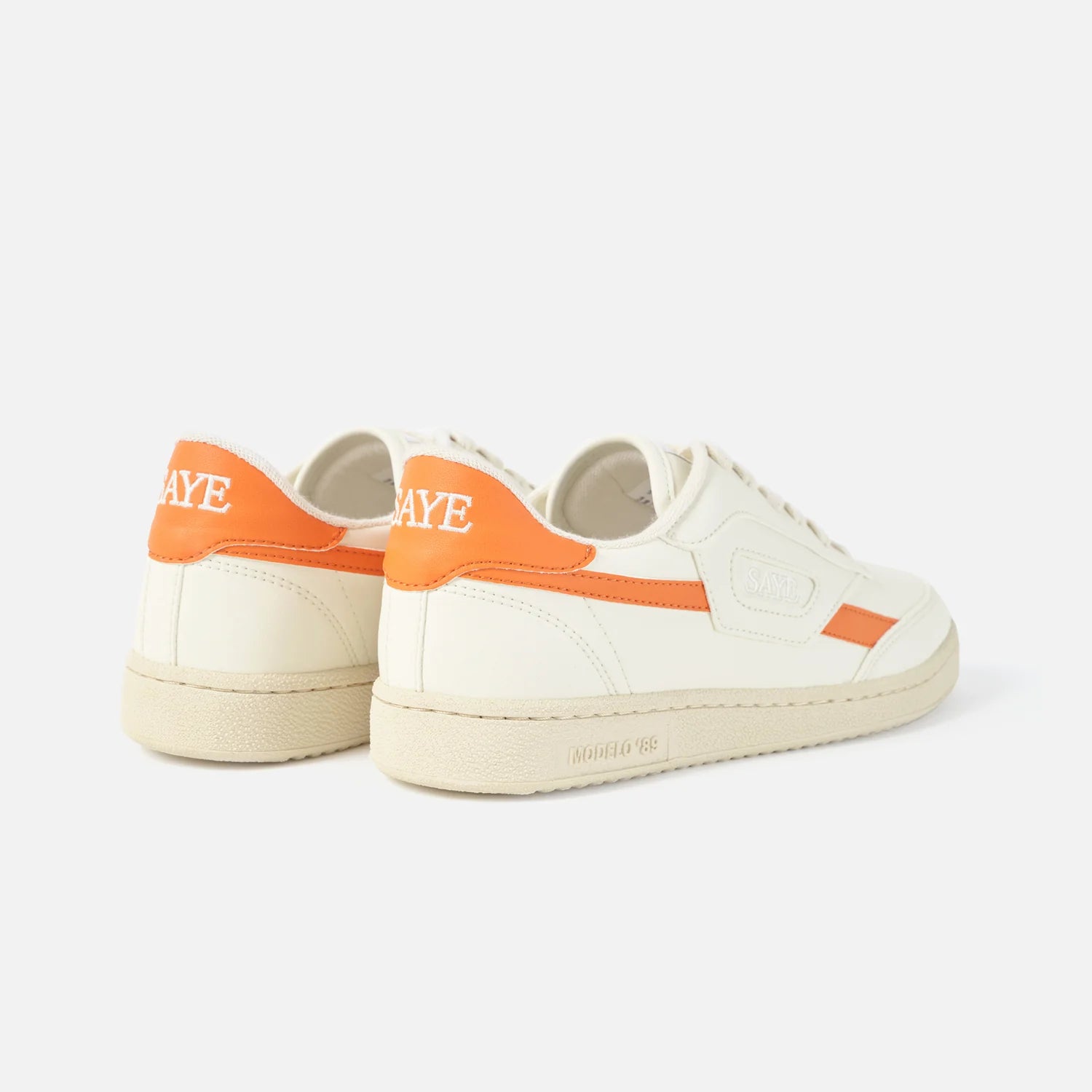 A pair of white SAYE vegan sneakers with orange accents and the text "Modelo '89" on the side.
