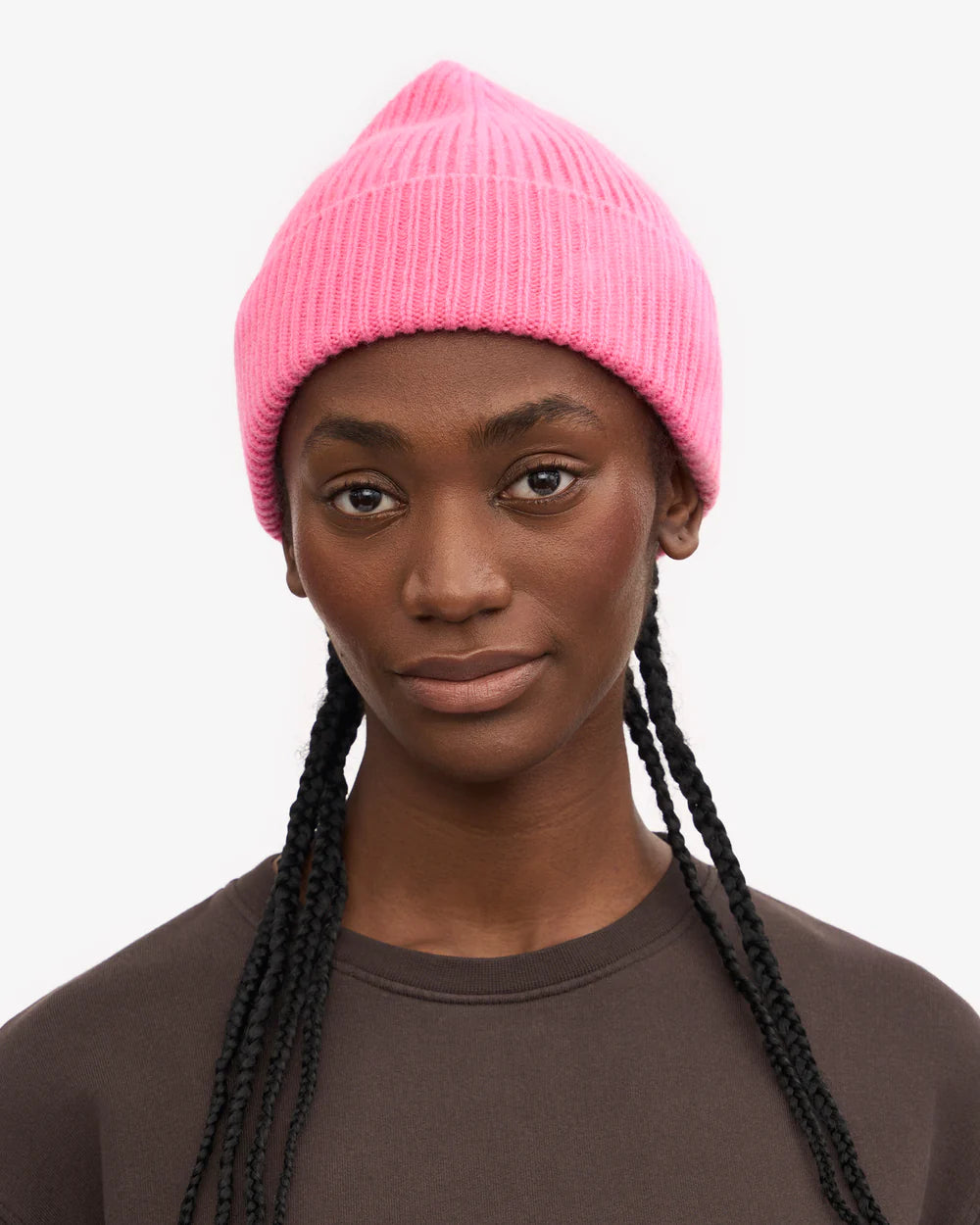 Person with long braids wears the Colorful Standard Merino Wool Beanie in bright pink, made from recycled merino wool, paired with a dark brown shirt, facing forward with a neutral expression against a plain backdrop.
