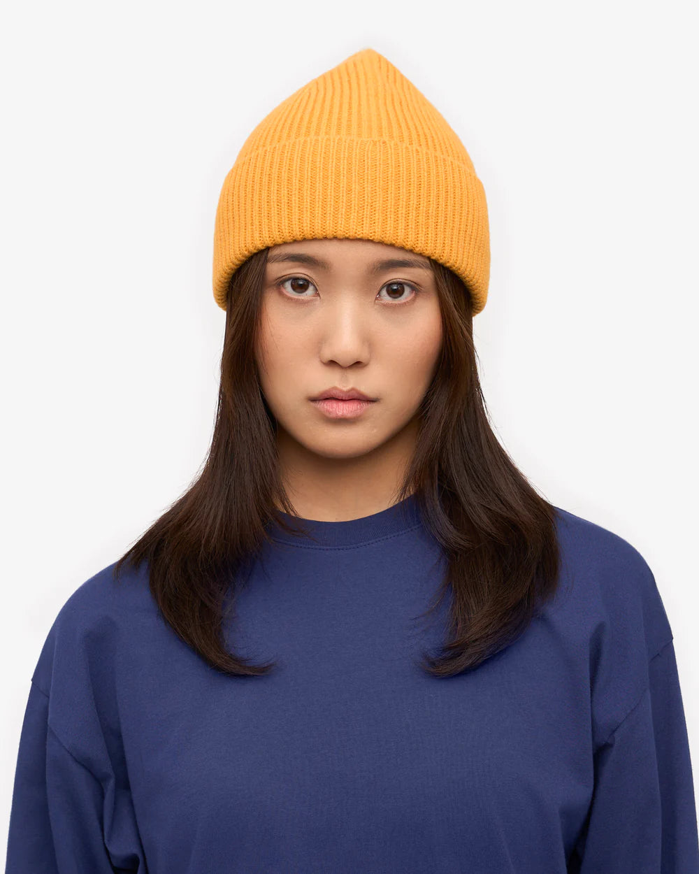 A person with long dark hair wears a yellow Colorful Standard Merino Wool Beanie and a blue shirt, looking directly at the camera against a plain white background.