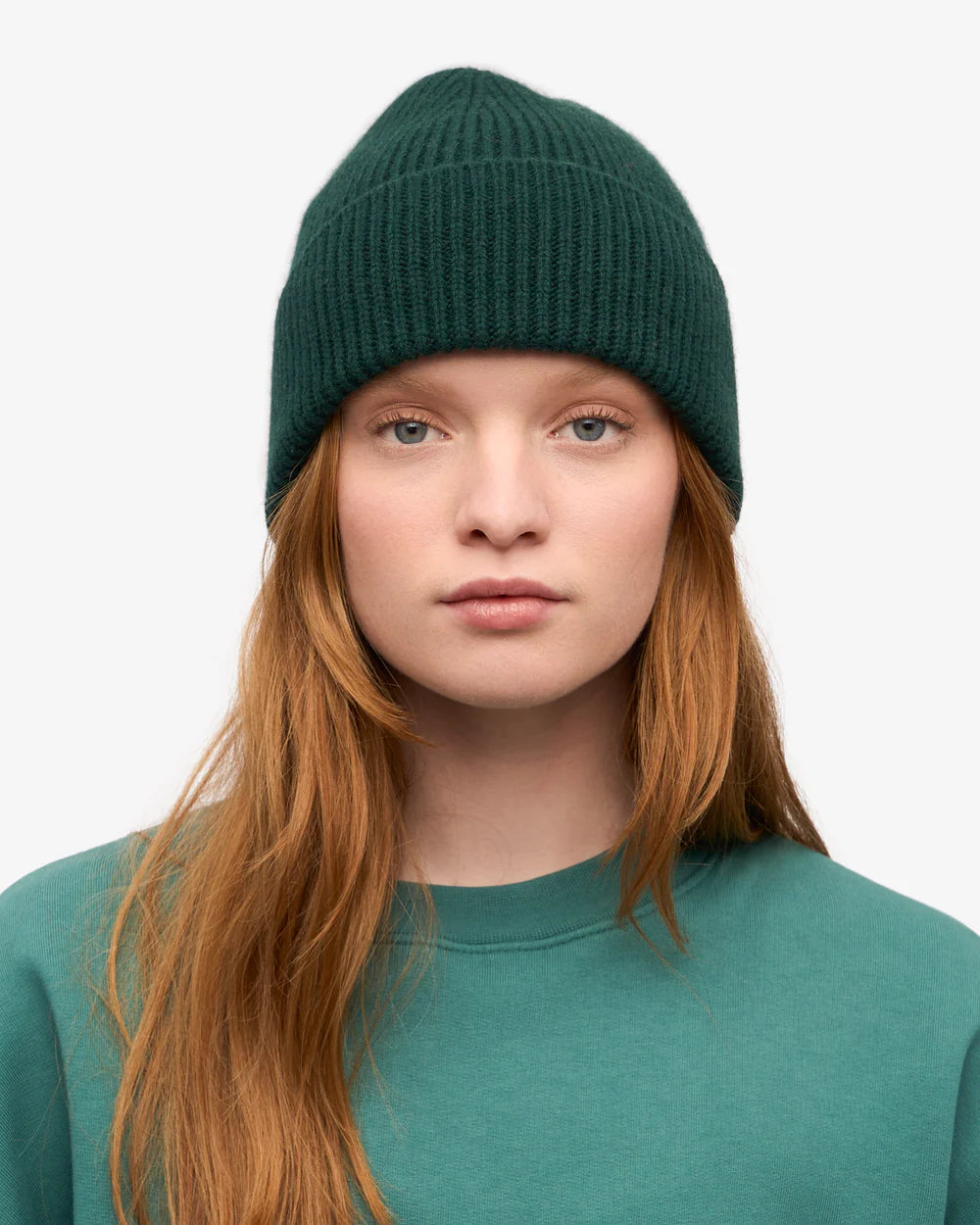A woman with long red hair faces the camera against a plain white background, wearing a dark green Colorful Standard Merino Wool Beanie made from recycled merino wool and a matching green sweatshirt.