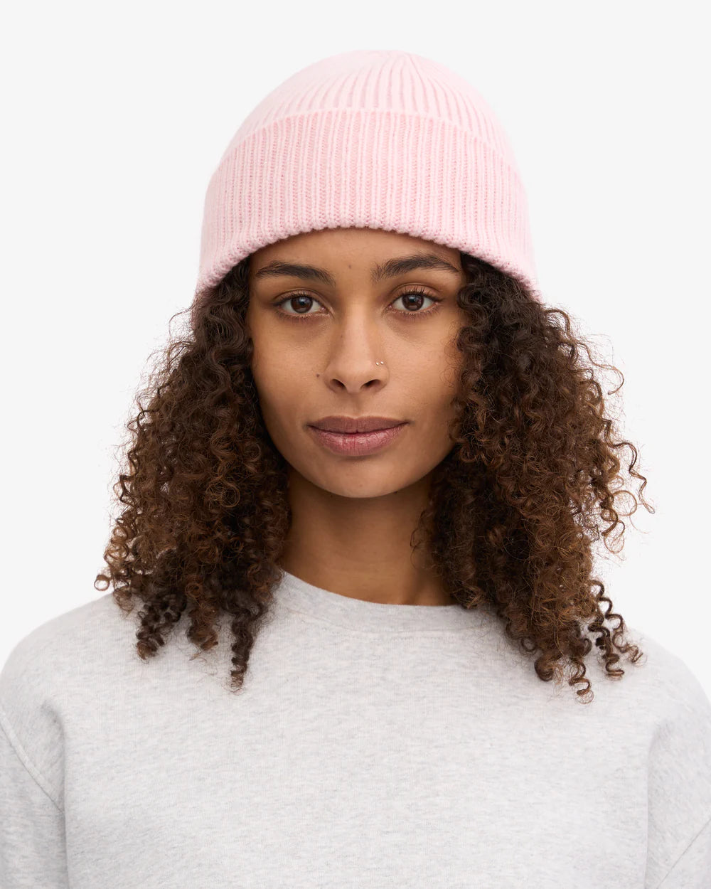 A woman with curly hair faces forward against a plain white background, wearing a light pink Colorful Standard Merino Wool Beanie and a light gray sweatshirt.