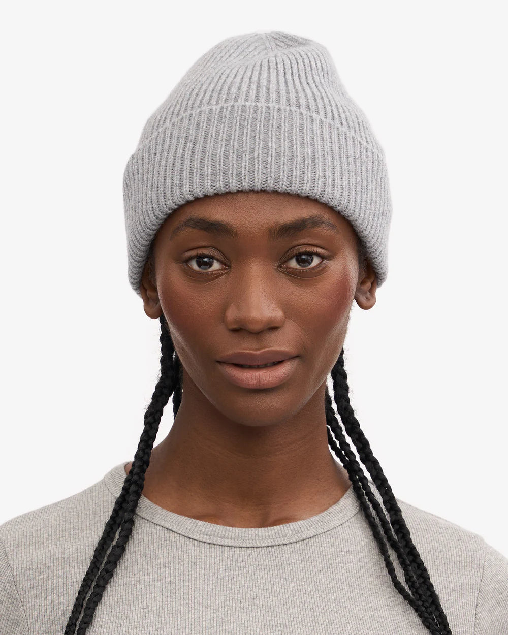 A person with long braids faces forward against a plain white background, wearing a light gray Merino Wool Beanie by Colorful Standard paired with a matching gray shirt.