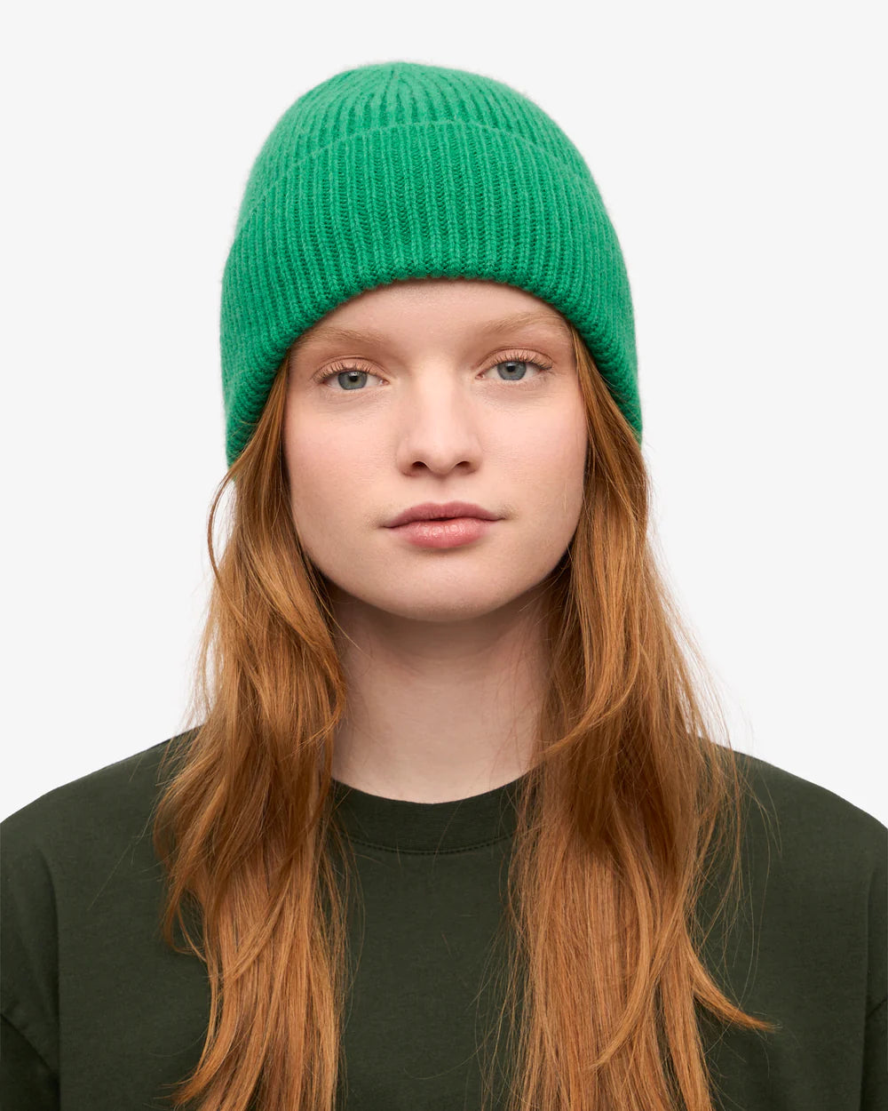 A person with long red hair wears a Colorful Standard Merino Wool Beanie in bright green and a dark green shirt, looking directly at the camera against a plain background.