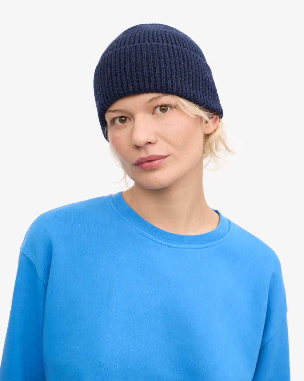 A person wearing a navy blue Merino Wool Beanie by Colorful Standard and a bright blue sweatshirt looks at the camera against a plain white background.