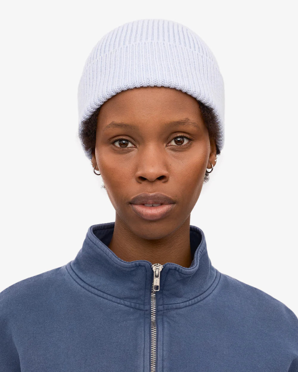 A person wearing a Colorful Standard Merino Wool Beanie in light blue, small hoop earrings, and a blue zip-up jacket, facing forward against a plain white background.