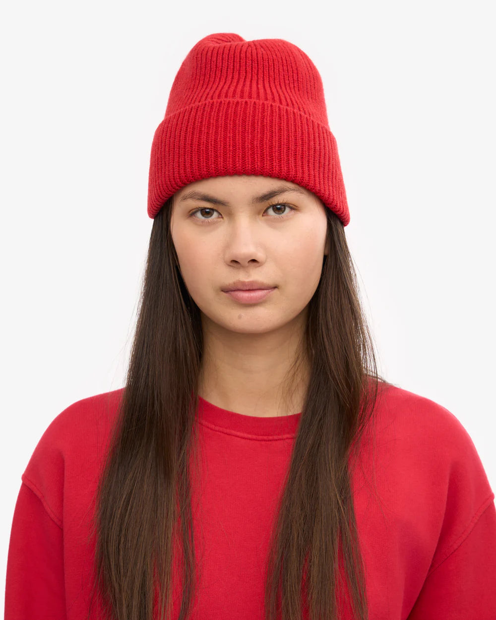 A person with long brown hair wears a red Colorful Standard Merino Wool Beanie and a red crewneck sweatshirt, facing the camera against a plain light background.