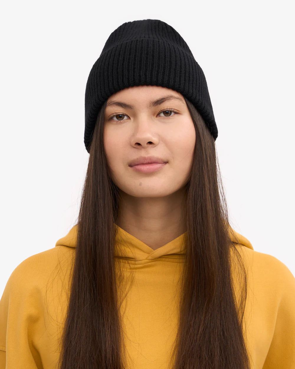 A person with long straight hair wears a Colorful Standard Merino Wool Beanie in black and a yellow hoodie, facing forward against a plain white background.