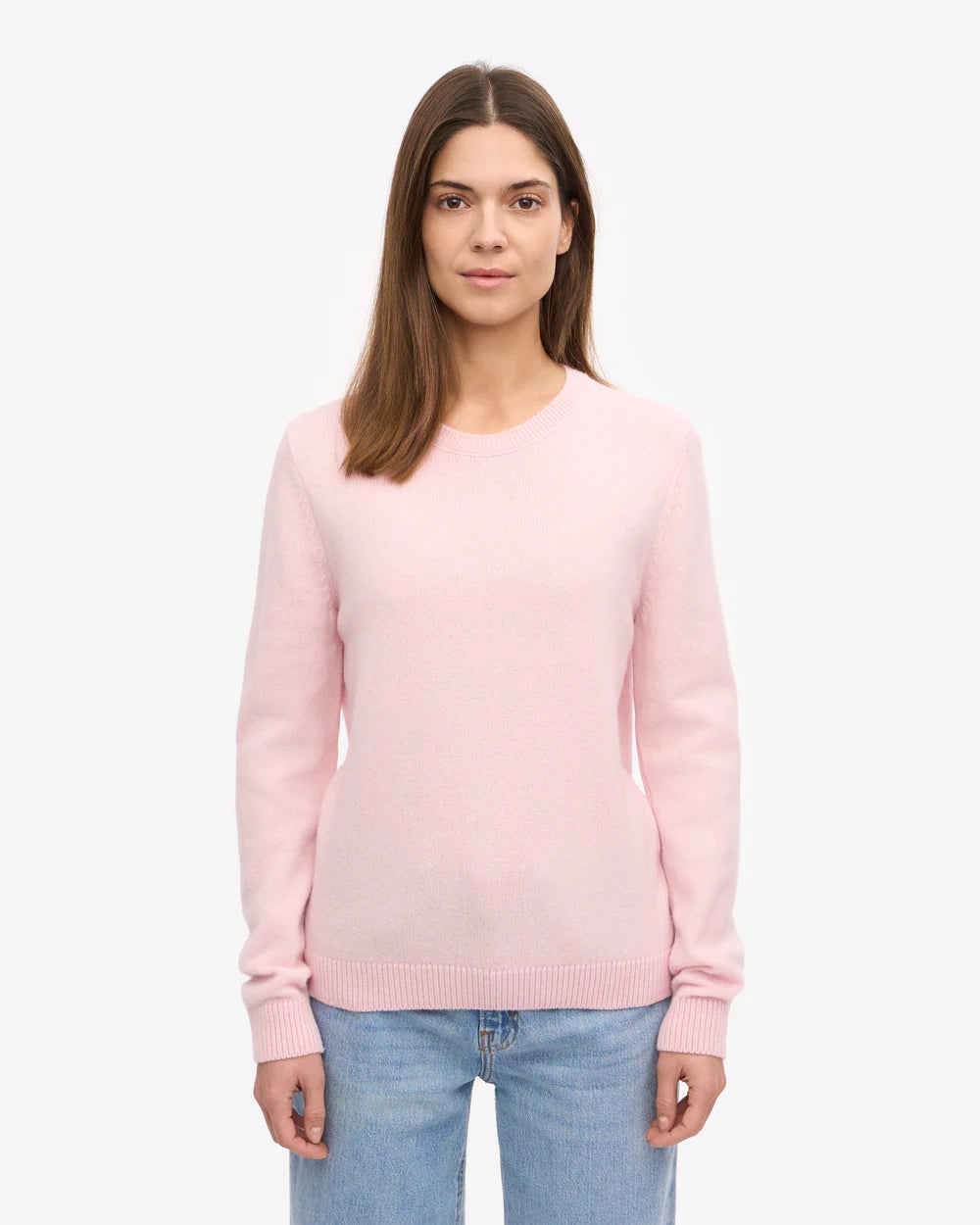 A woman with straight brown hair wears the Colorful Standard Classic Merino Wool Crew in Faded Pink with blue jeans, standing against a plain white background.