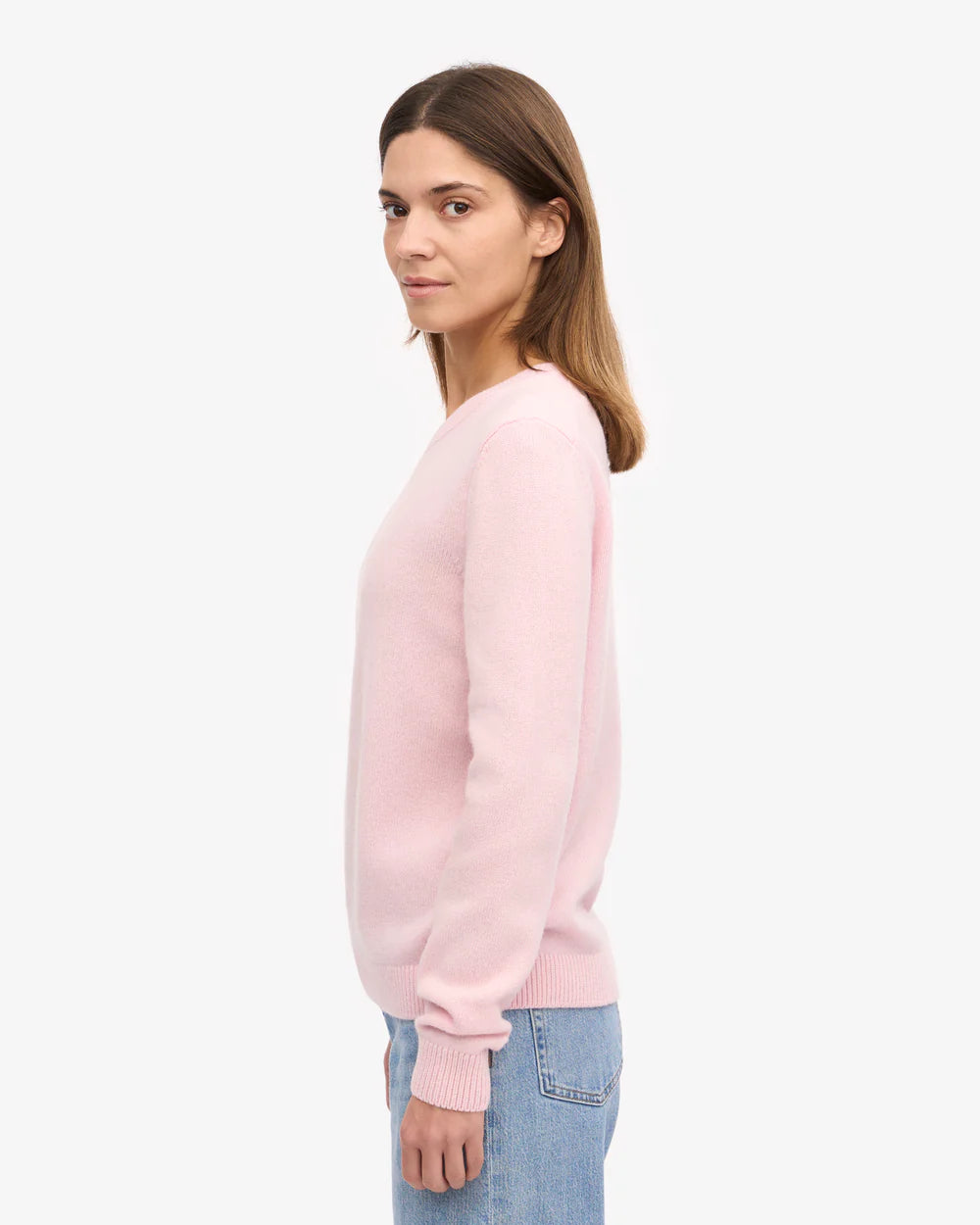 A person stands in profile wearing the Colorful Standard Classic Merino Wool Crew in Faded Pink and light blue jeans against a plain white background.