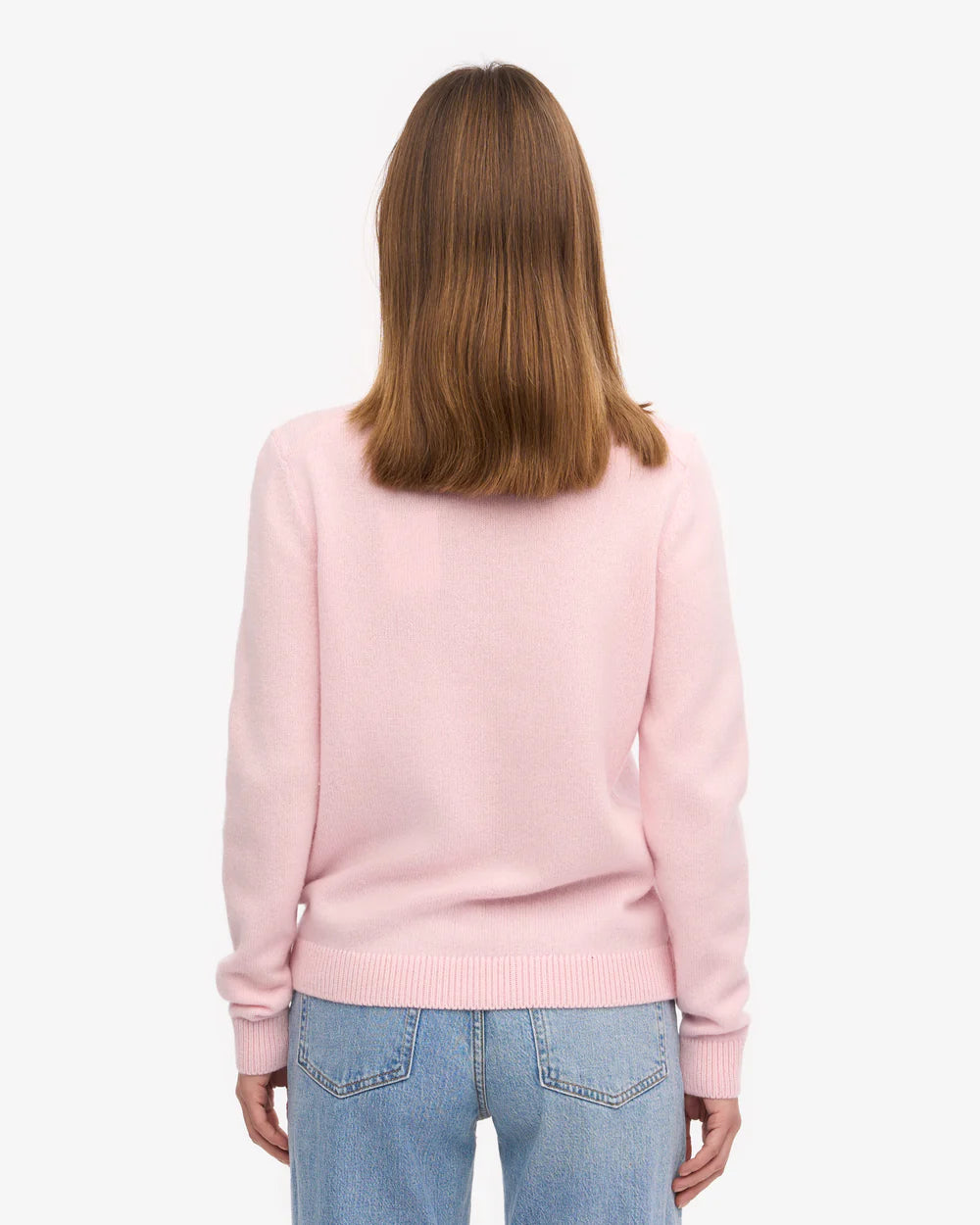 A person with shoulder-length brown hair is seen from the back wearing the Colorful Standard Classic Merino Wool Crew in Faded Pink and light blue jeans, set against a plain white background.