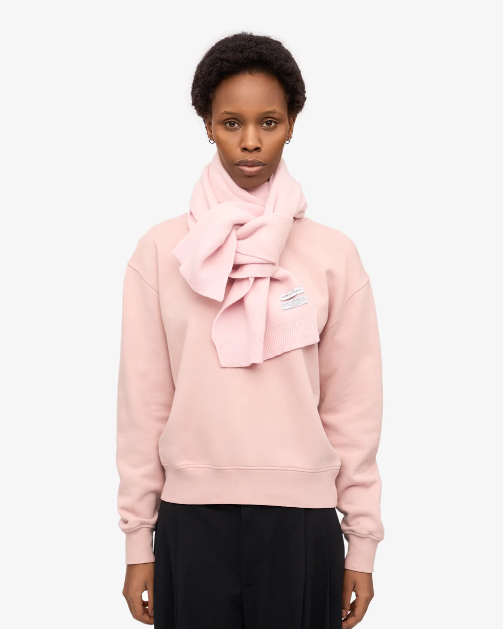 A person faces forward against a plain white background, wearing a pale pink sweatshirt and the Colorful Standard Merino Wool Scarf.