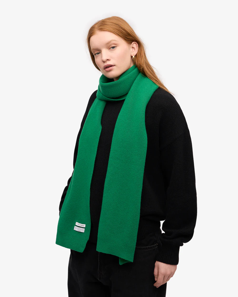 A person with long red hair wears a black sweater and the bright green Merino Wool Scarf by Colorful Standard, standing against a plain light background.