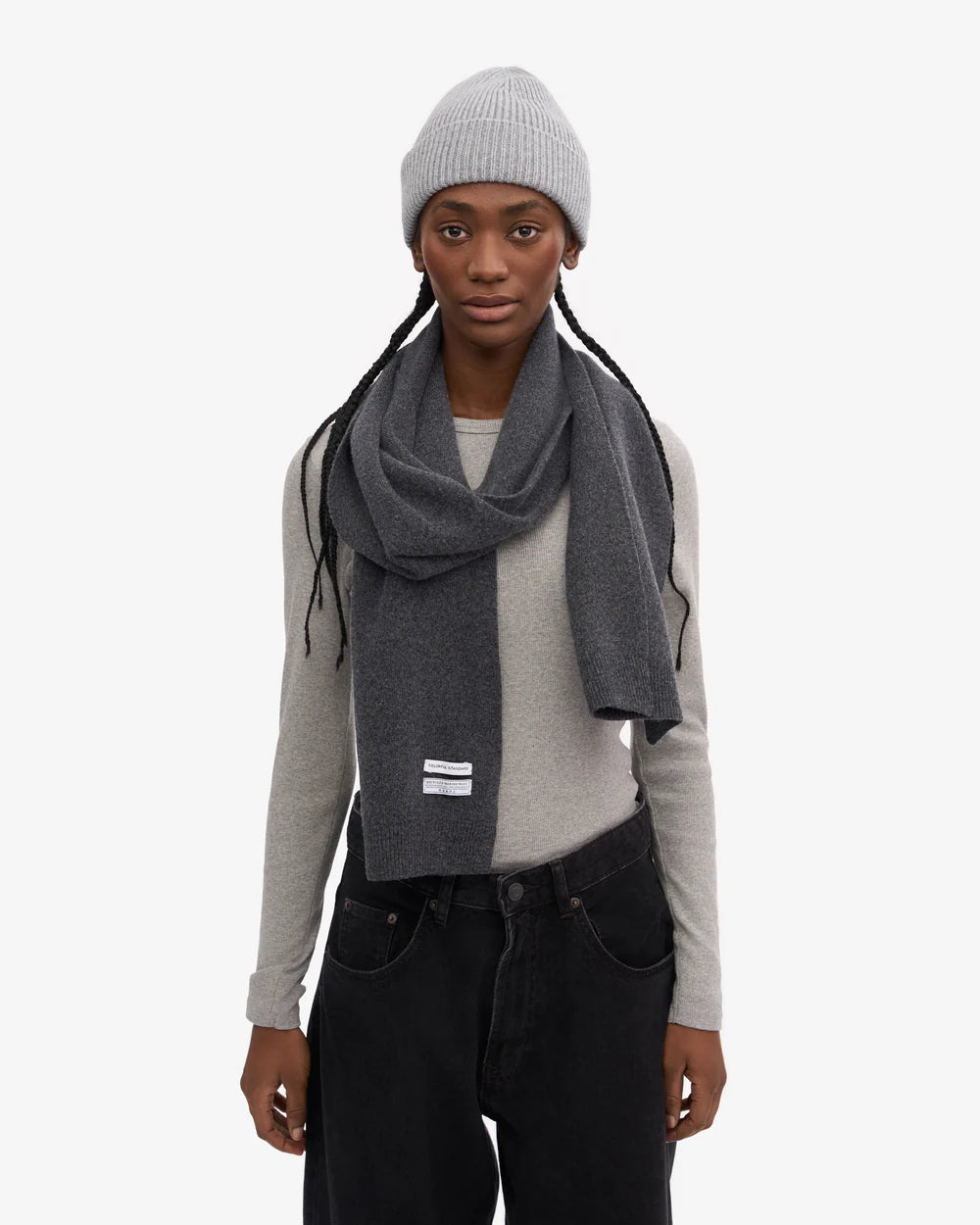 A person stands against a plain white background wearing a gray beanie, Colorful Standard Merino Wool Scarf, light gray long-sleeve shirt, and black pants.
