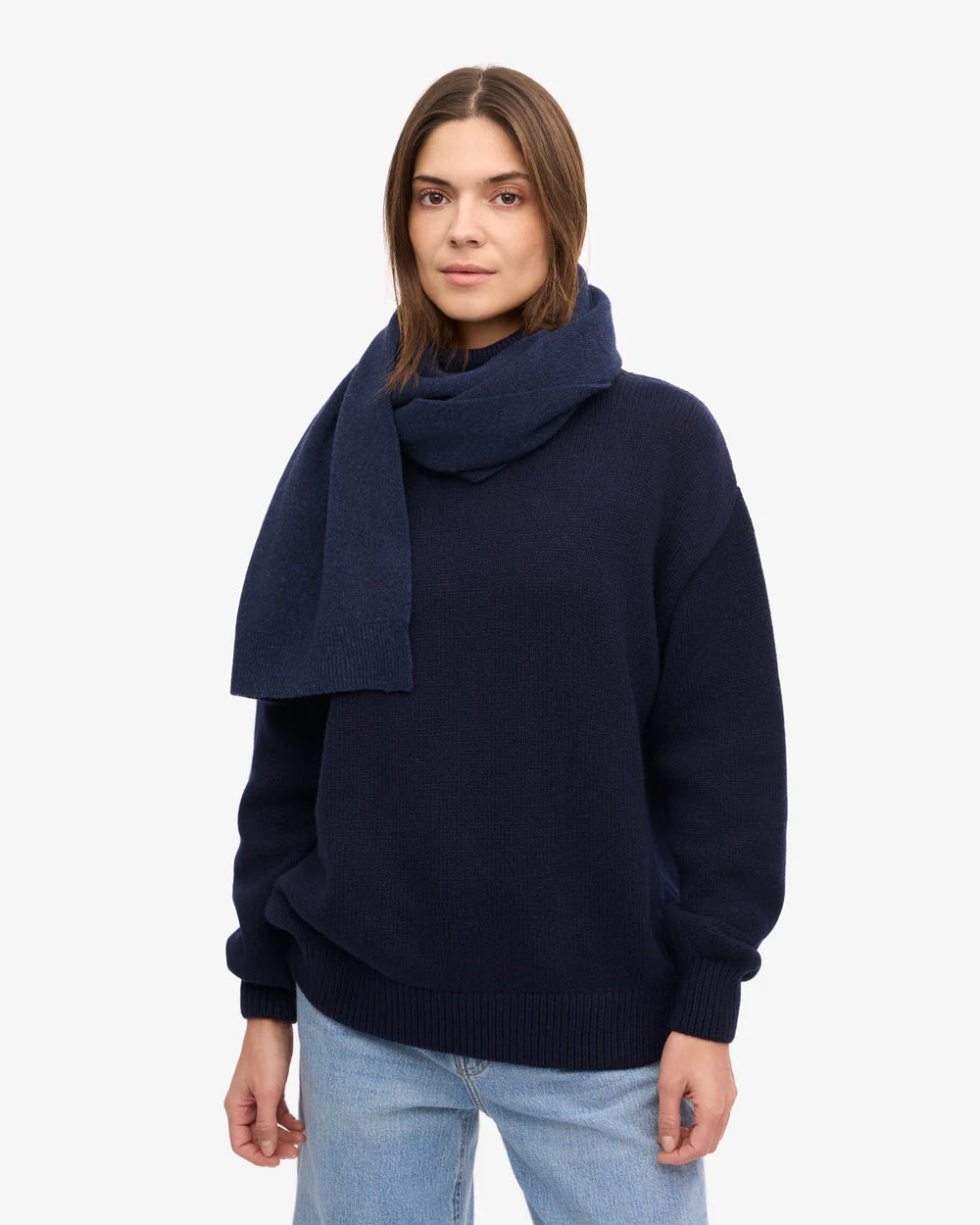 A person stands facing forward against a white background, wearing light blue jeans, a navy sweater, and the Colorful Standard Merino Wool Scarf, with a neutral expression.