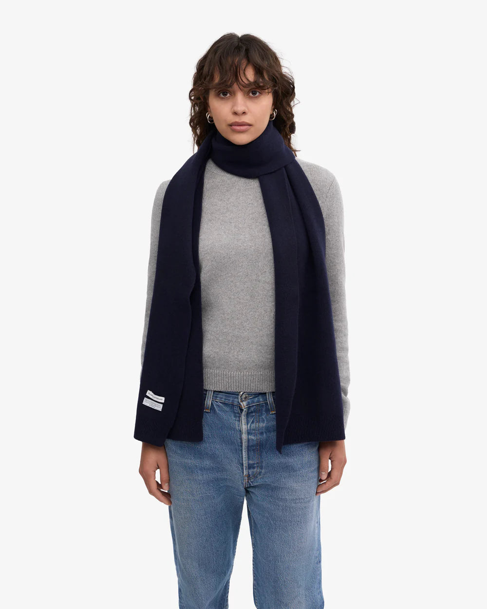 A woman stands against a plain white background wearing a gray sweater, blue jeans, and the Colorful Standard Merino Wool Scarf.