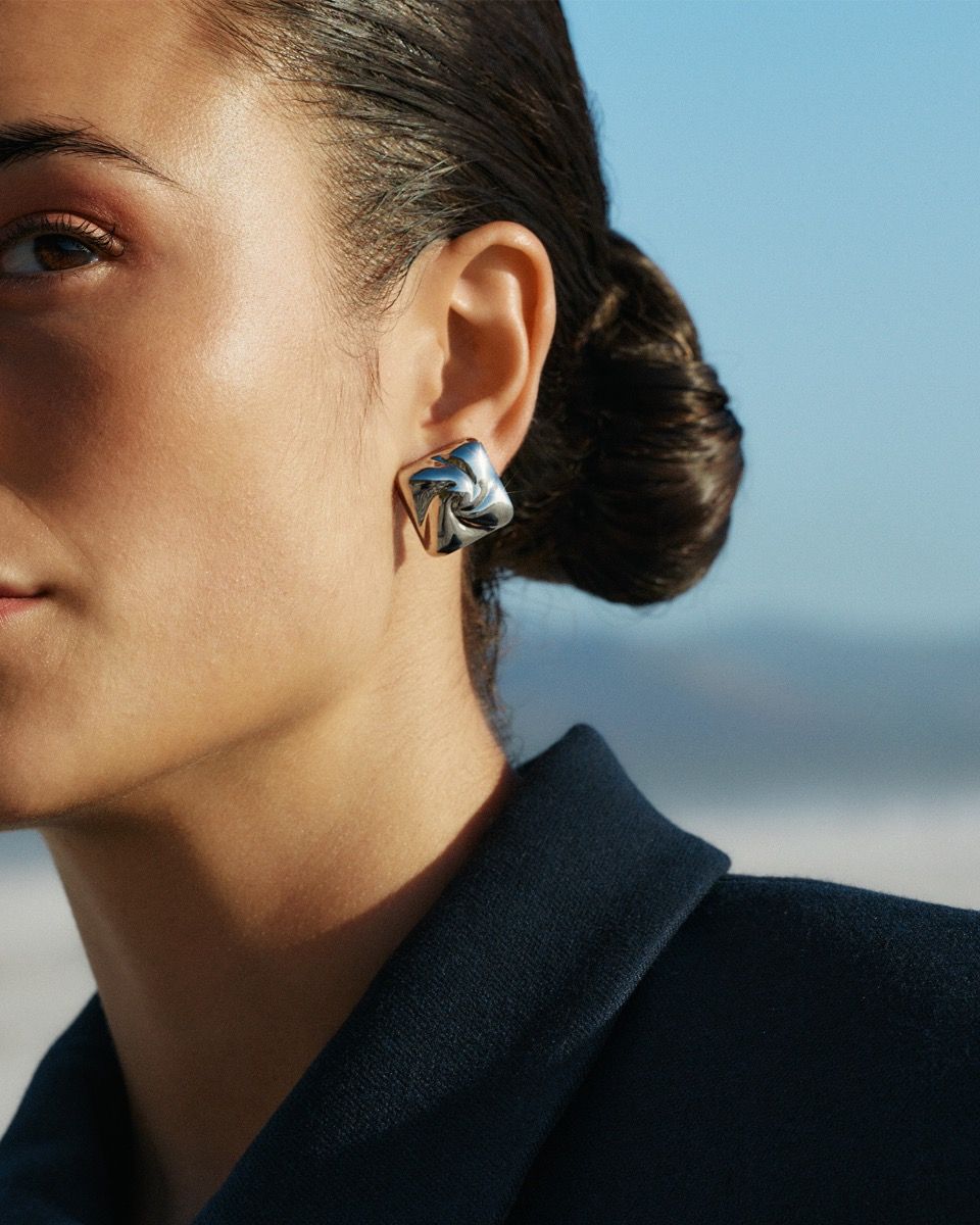 Close-up of a person wearing EDBLAD Milky Way Studs S earrings and a dark blazer with hair tied back, set against a blurred outdoor background.