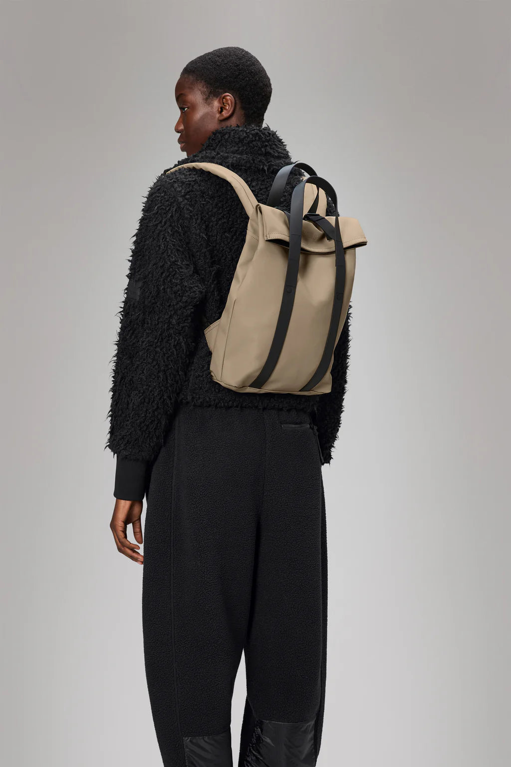 A person in a black textured jacket and pants stands sideways, showcasing the Rains 2 Way Tote Backpack Mini—a commuter-friendly beige roll-top bag with black straps—against a plain background.