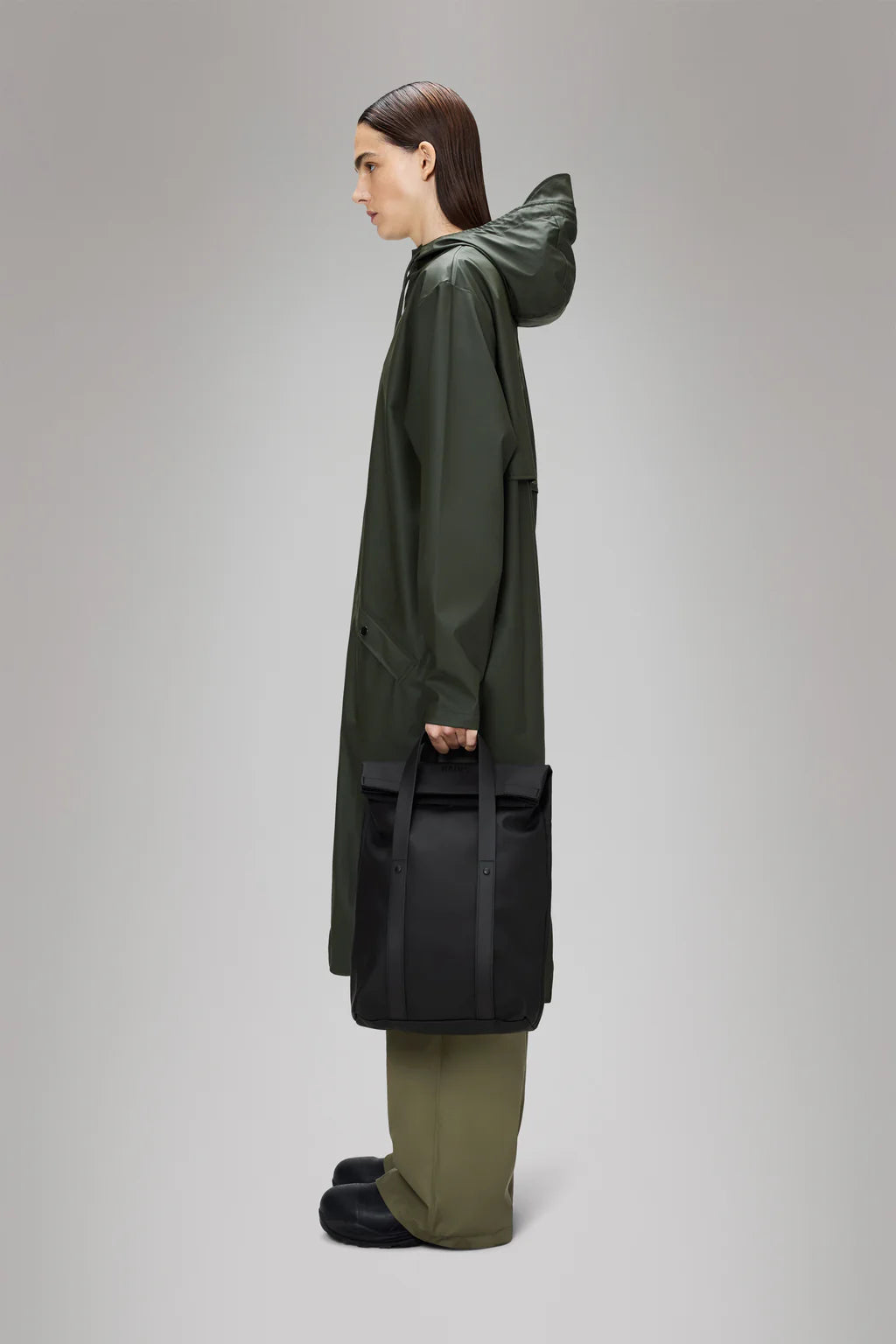 Person in a long, olive green raincoat and green pants stands in profile holding the Rains 2 Way Tote Backpack Mini, a commuter-friendly black bag, against a plain gray background.
