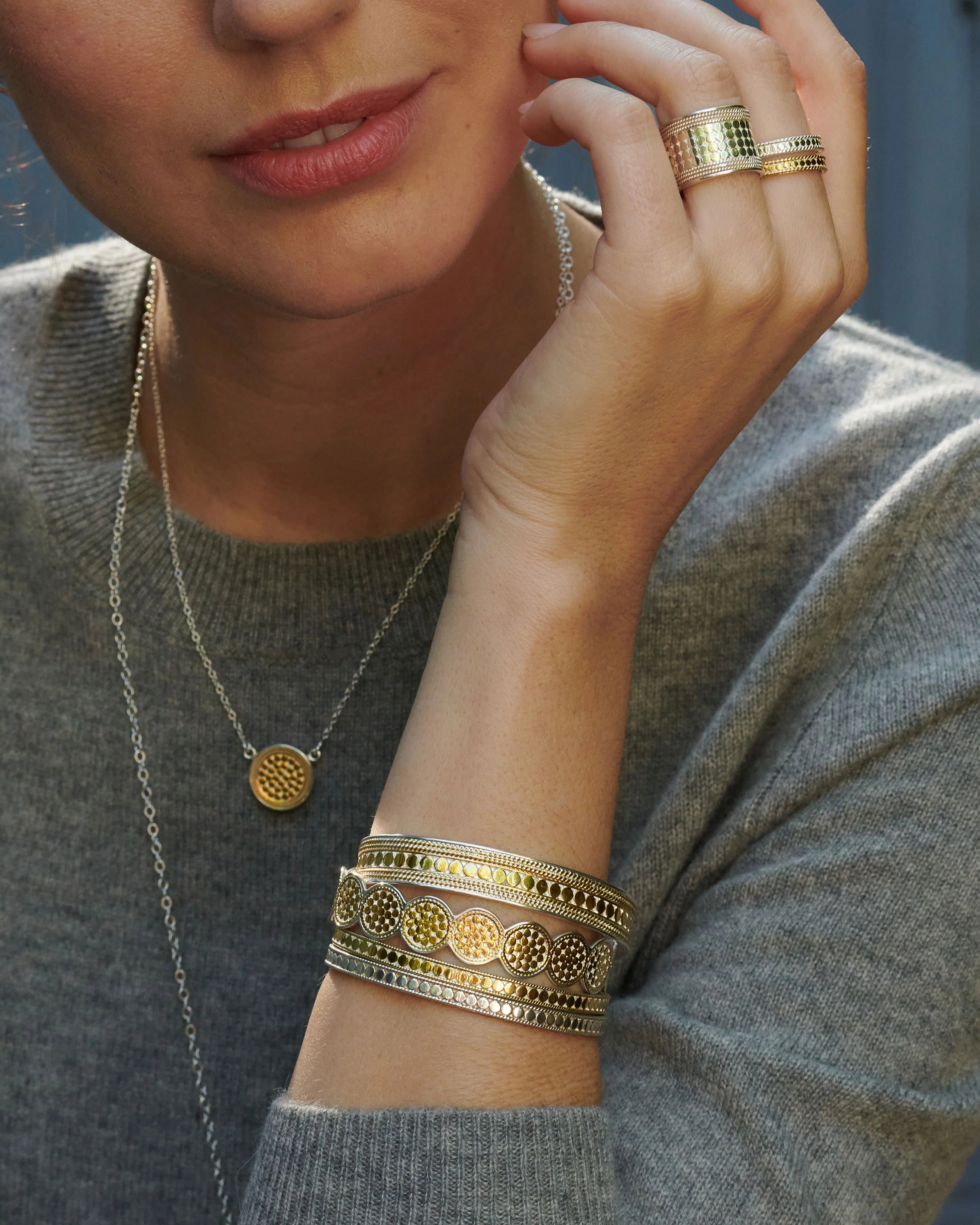 A person wearing a gray sweater showcases jewelry, including two Classic Multi-Disc Cuffs in 18k gold plating by Anna Beck, a gold ring, and a gold necklace with a round pendant.