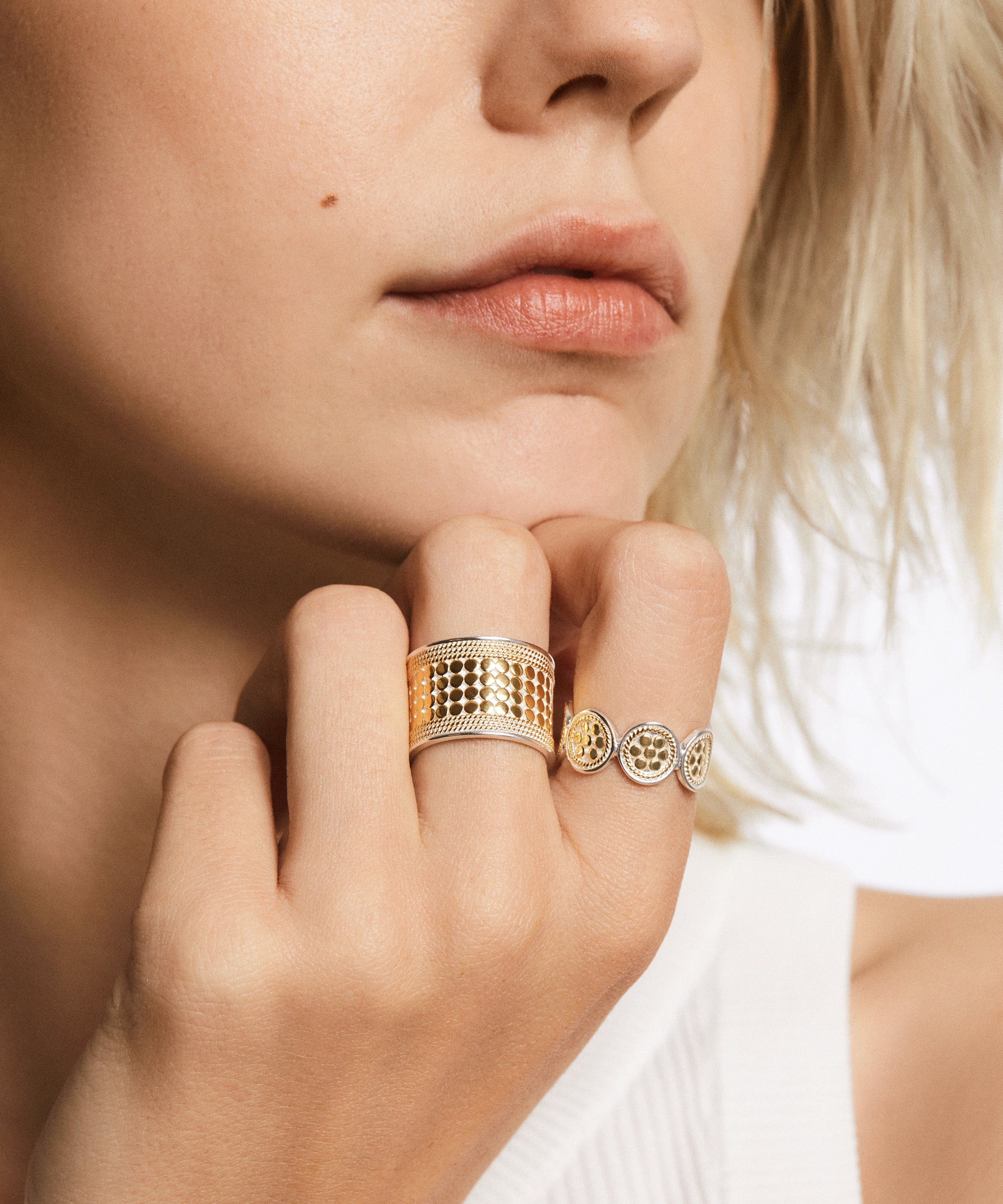 A person with light skin and short blond hair wears two gold rings on their hand, one featuring a wide band with an intricate design symbolizing an endless connection and the other being the Anna Beck Classic Multi-Disc Ring - Gold, adorned with three circular elements.