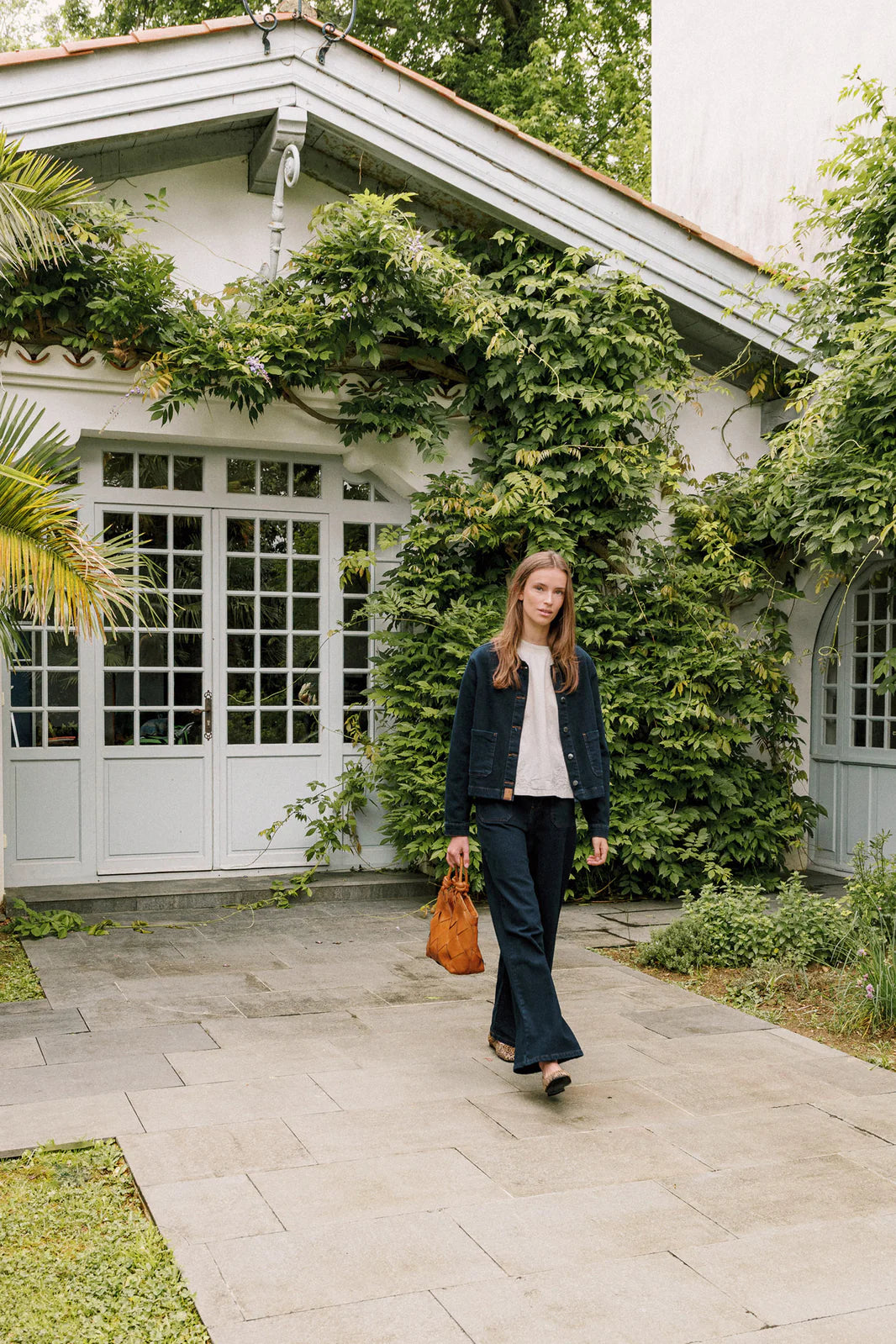 A woman in Indi & Cold Wide Leg Denim Trousers - Navy and a denim jacket with patch pockets walks on a stone path by a house with large windows and greenery, carrying a brown bag.