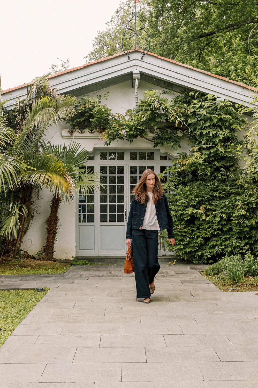 A woman in a dark jacket and Indi & Cold’s Wide Leg Denim Trousers - Navy walks on a stone path toward the camera, with a house, glass doors, and lush green plants in the background.