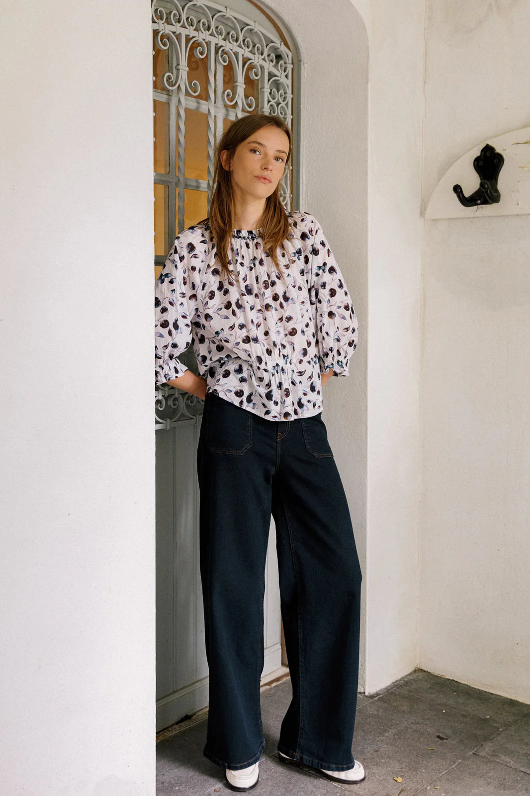 A woman stands in a doorway wearing a white patterned blouse, Indi & Cold Wide Leg Denim Trousers in navy with patch pockets, and white shoes, her hands tucked in her pockets.