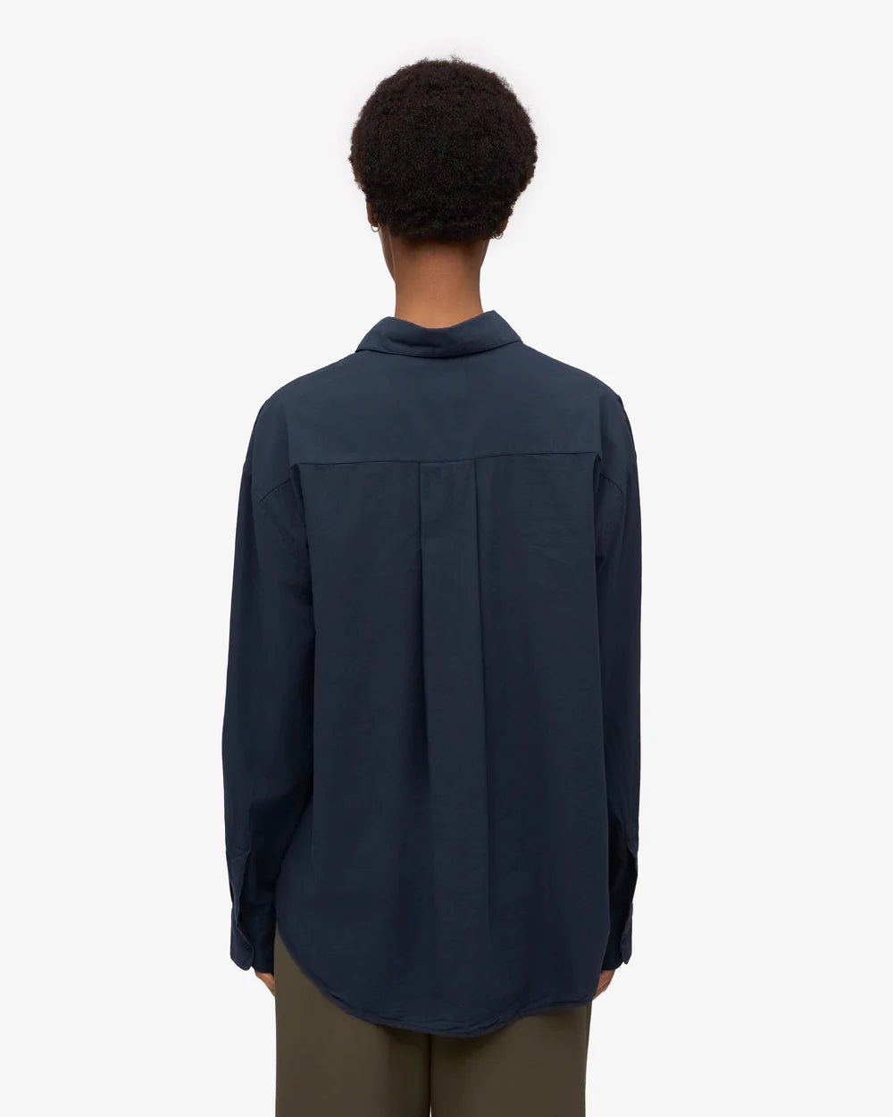 A person with short curly hair is seen from behind, wearing the Colorful Standard Organic Oversized Shirt in navy blue and olive green pants against a plain light background.