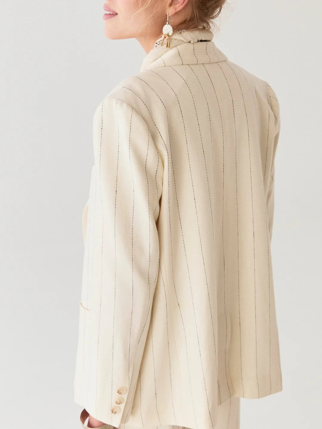 A woman is seen from the side in Maison Hotel's Paddy Jacket, featuring ecru stripes, paired with elegant earrings.