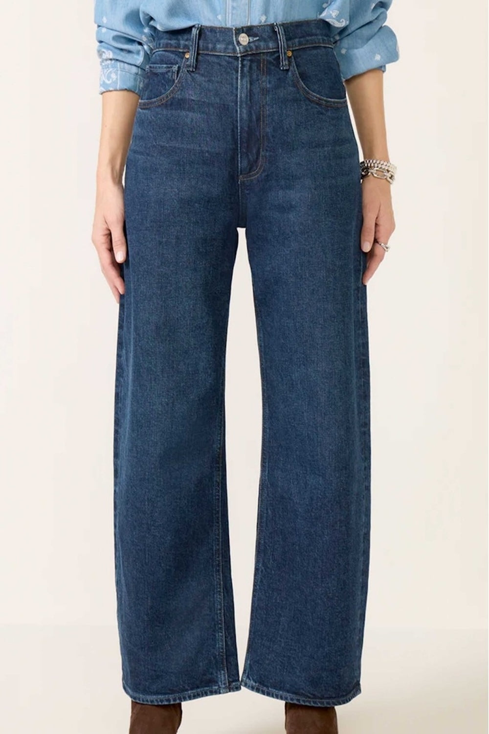 A person wears Paige's Arellia Skimmer Barrel Leg - Visage jeans in a dark vintage-inspired wash with a light blue shirt; hands at sides and brown shoes partially visible.