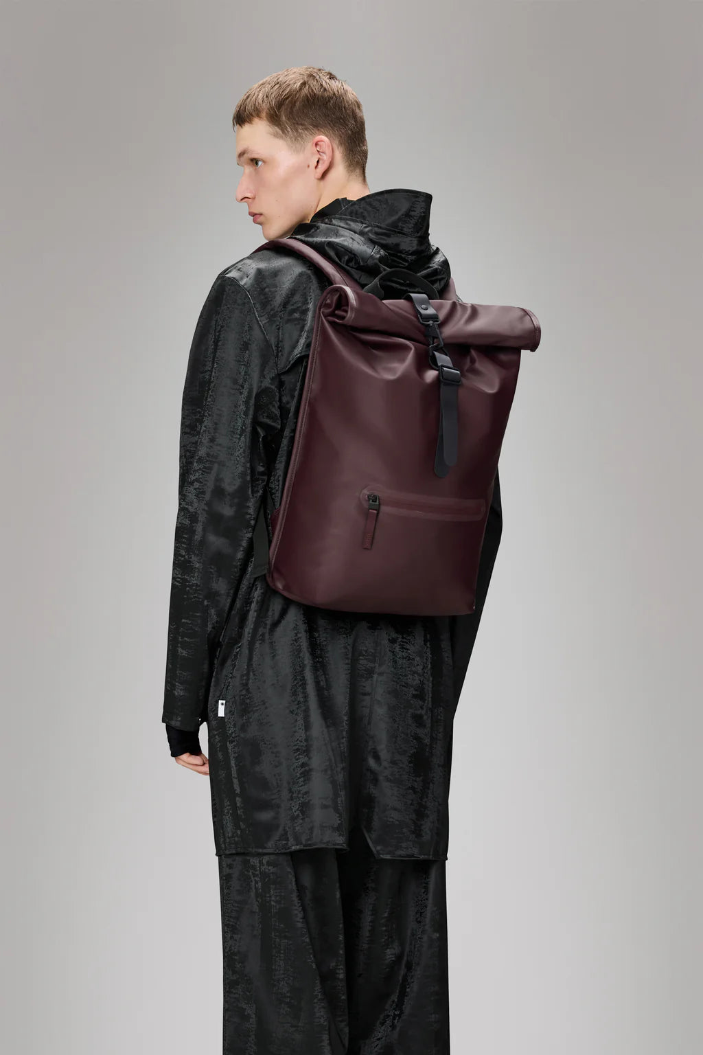 A person in a black textured jacket and pants stands facing away, highlighting the large burgundy Rolltop Rucksack - Depth by Rains—an ideal waterproof backpack for bike commutes.