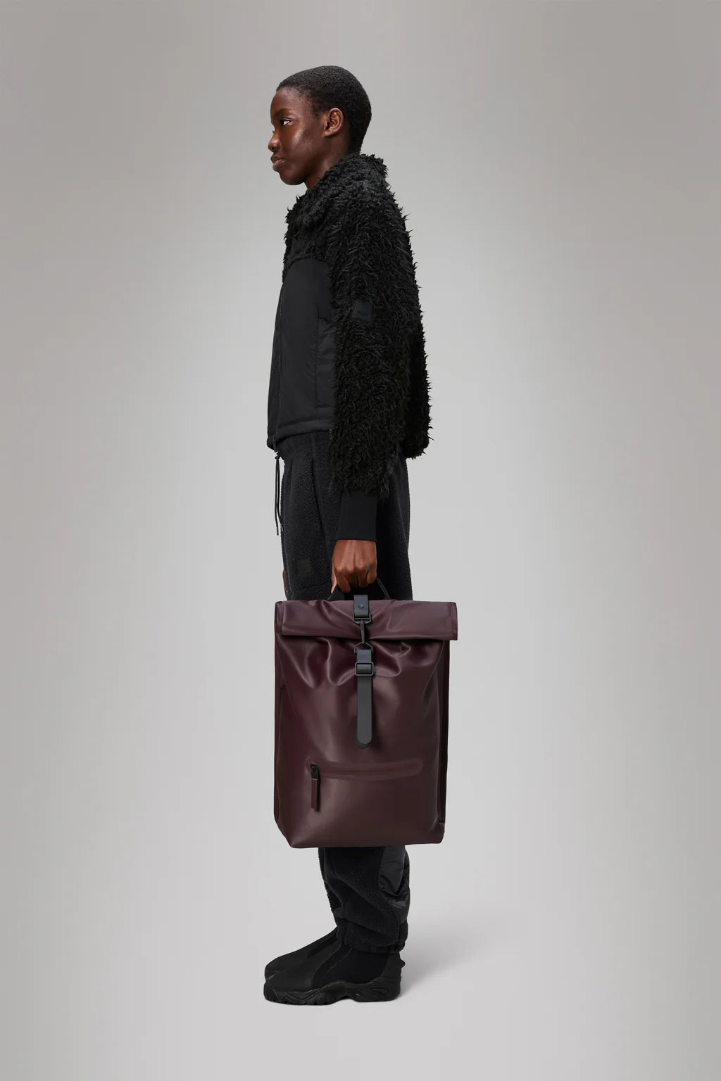 A person in profile holds the Rolltop Rucksack - Depth by Rains—a burgundy commuter bag—dressed in black pants, textured black jacket, and black shoes, set against a plain light gray background.