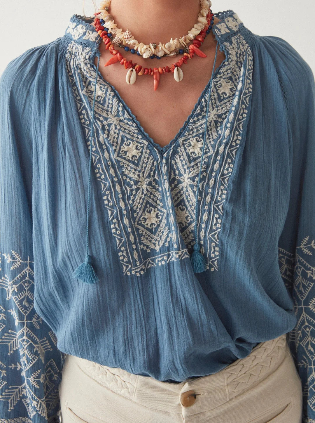 Close-up of a person wearing a Sandrine Cotton Blouse in French Blue tone with ethnic pattern embroidery and a beaded necklace by Maison Hotel.