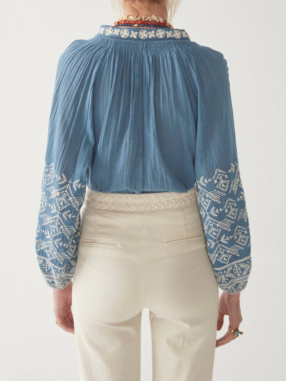 A woman from behind wearing a Sandrine Cotton Blouse in French Blue by Maison Hotel with floral embroidery and lace sleeve details and cream trousers.