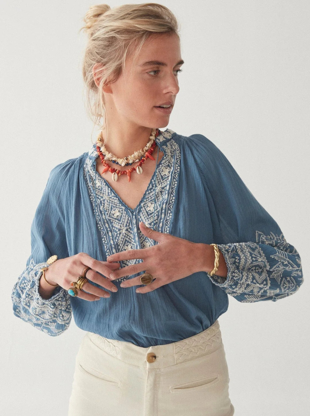 A woman in a Sandrine Cotton Blouse - French Blue from Maison Hotel and white pants accessorized with a red necklace and multiple rings looking to the side.